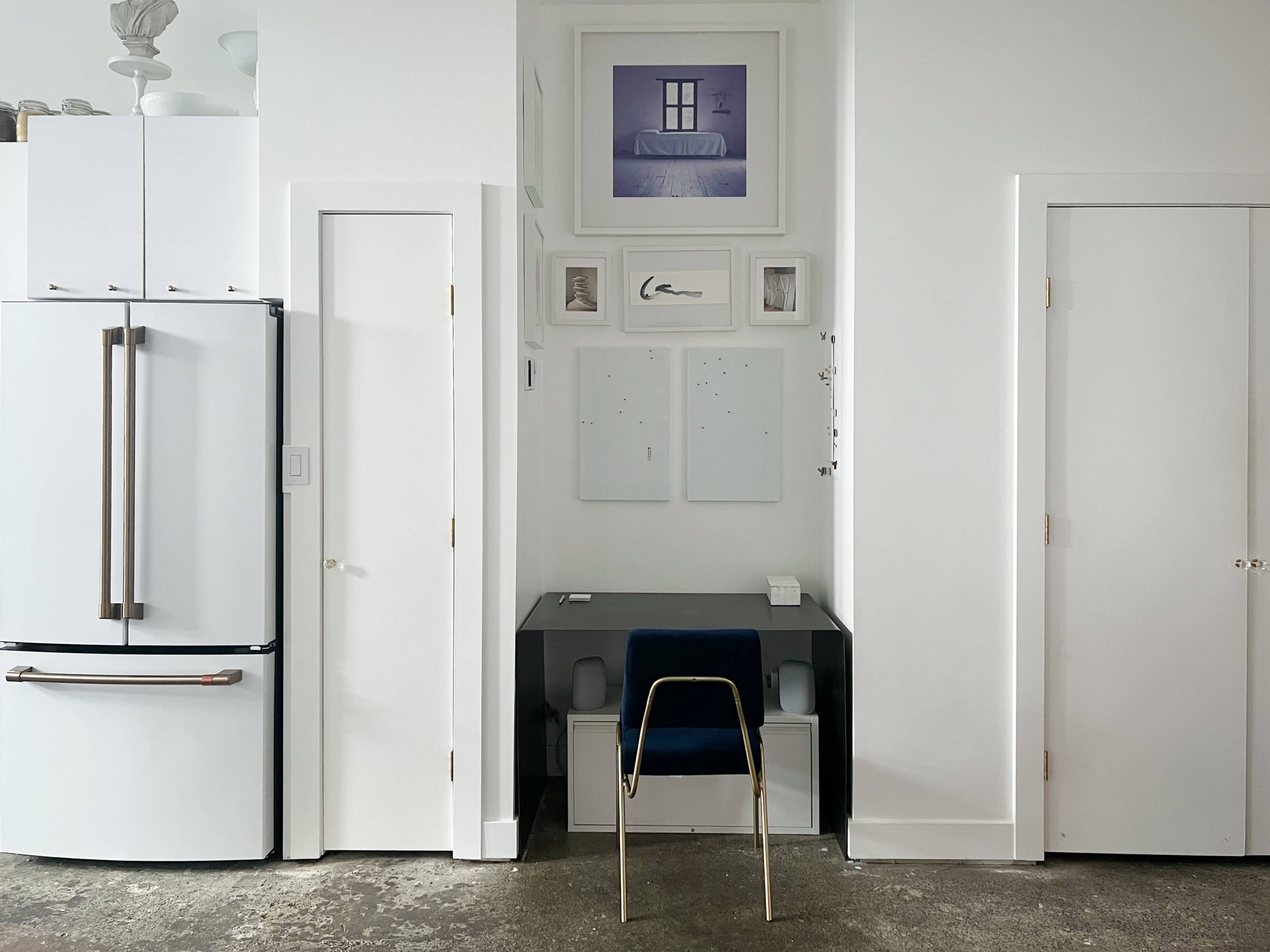 A minimalist kitchen and workspace area with a black desk and a chair positioned against a white wall, flanked by a refrigerator and two doors.