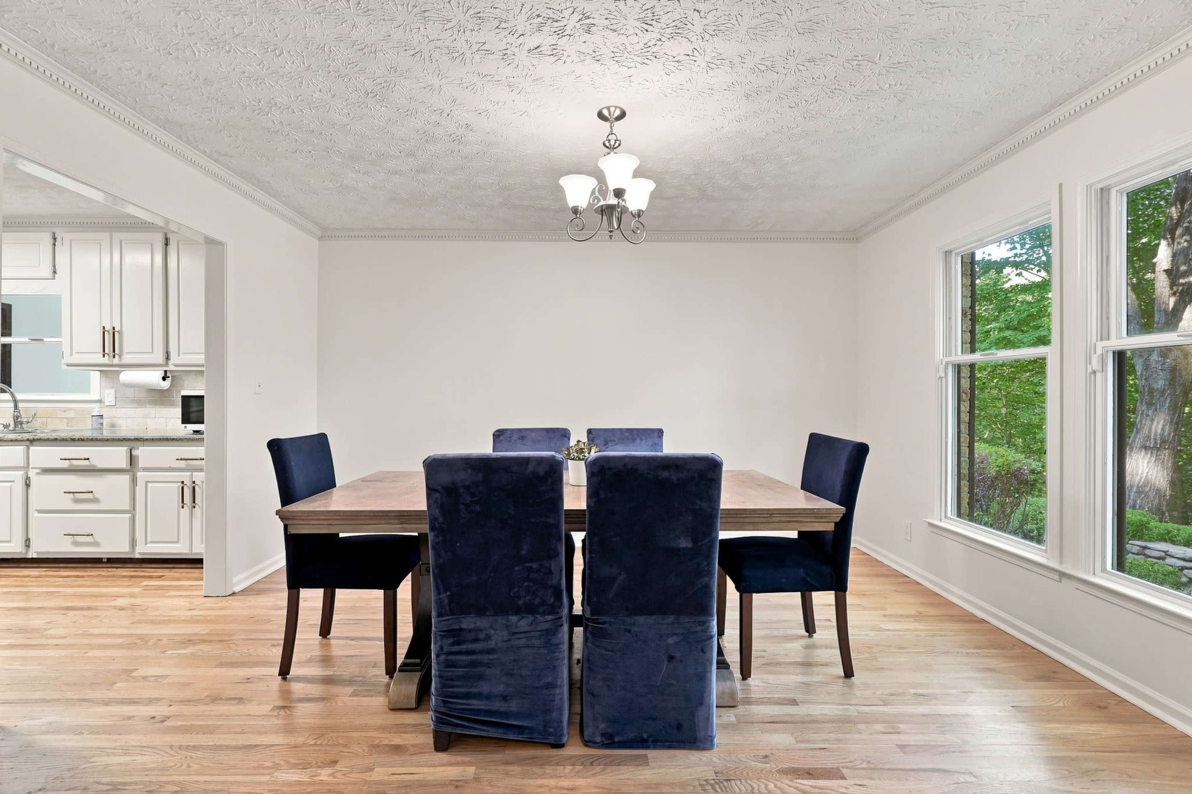 The image shows a spacious dining room with a wooden table surrounded by six dark blue upholstered chairs and two large windows allowing natural light to enter.