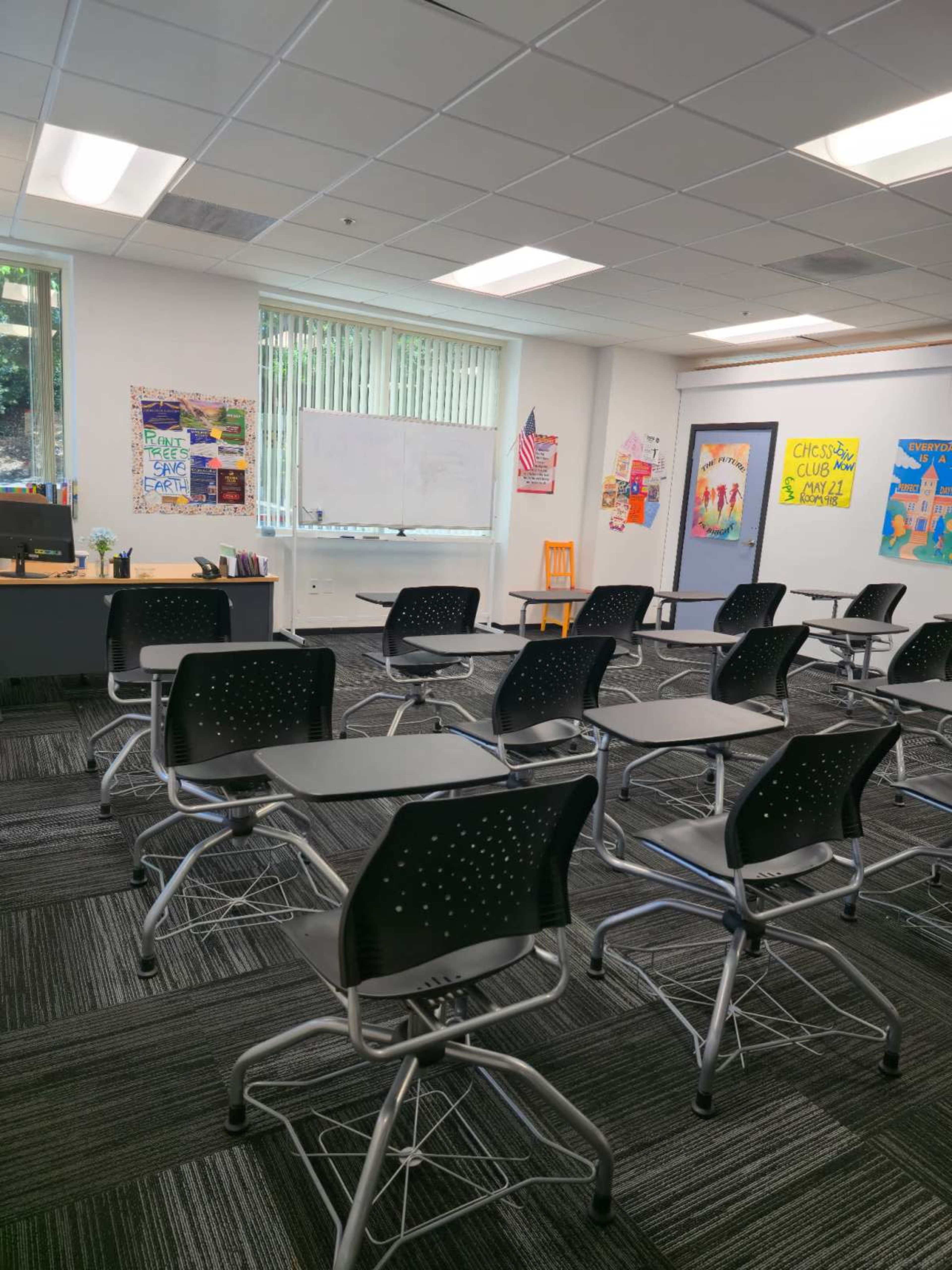 The image shows a neatly arranged classroom with several black desks facing the front, a whiteboard, and educational posters on the walls.