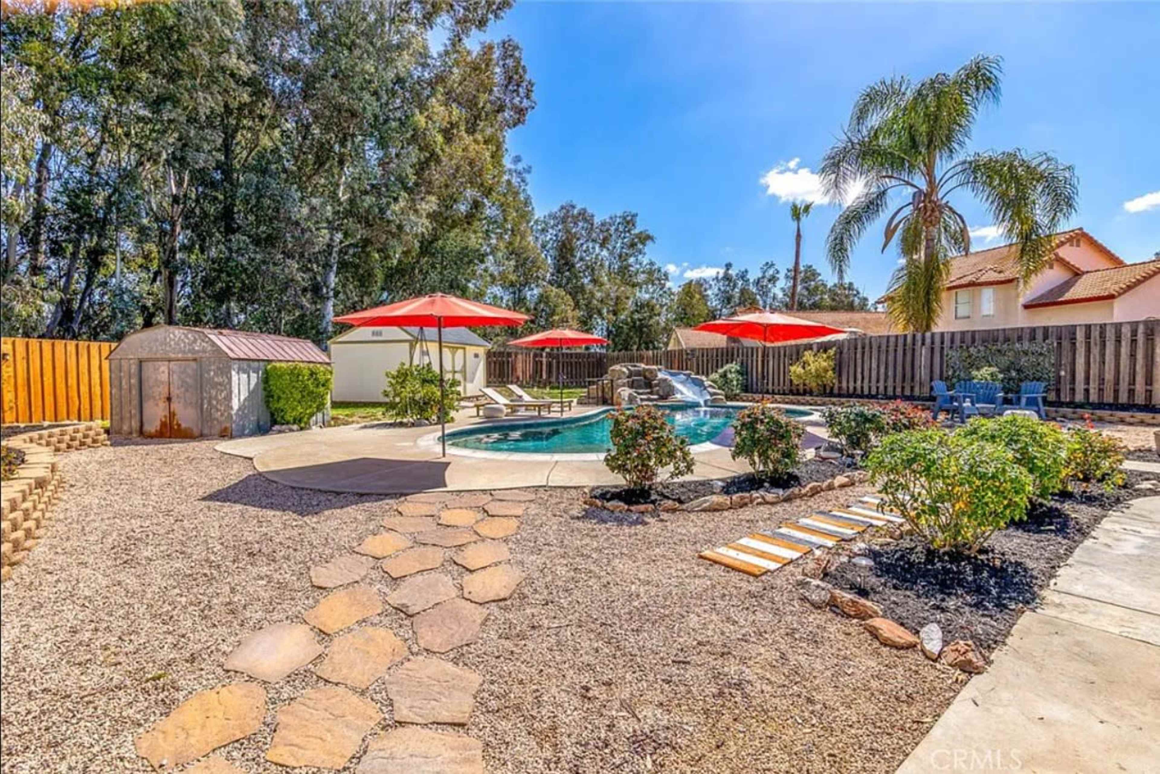 A landscaped backyard with a swimming pool, surrounded by stone pathways, patio furniture, and red umbrellas, along with a storage shed and various trees.