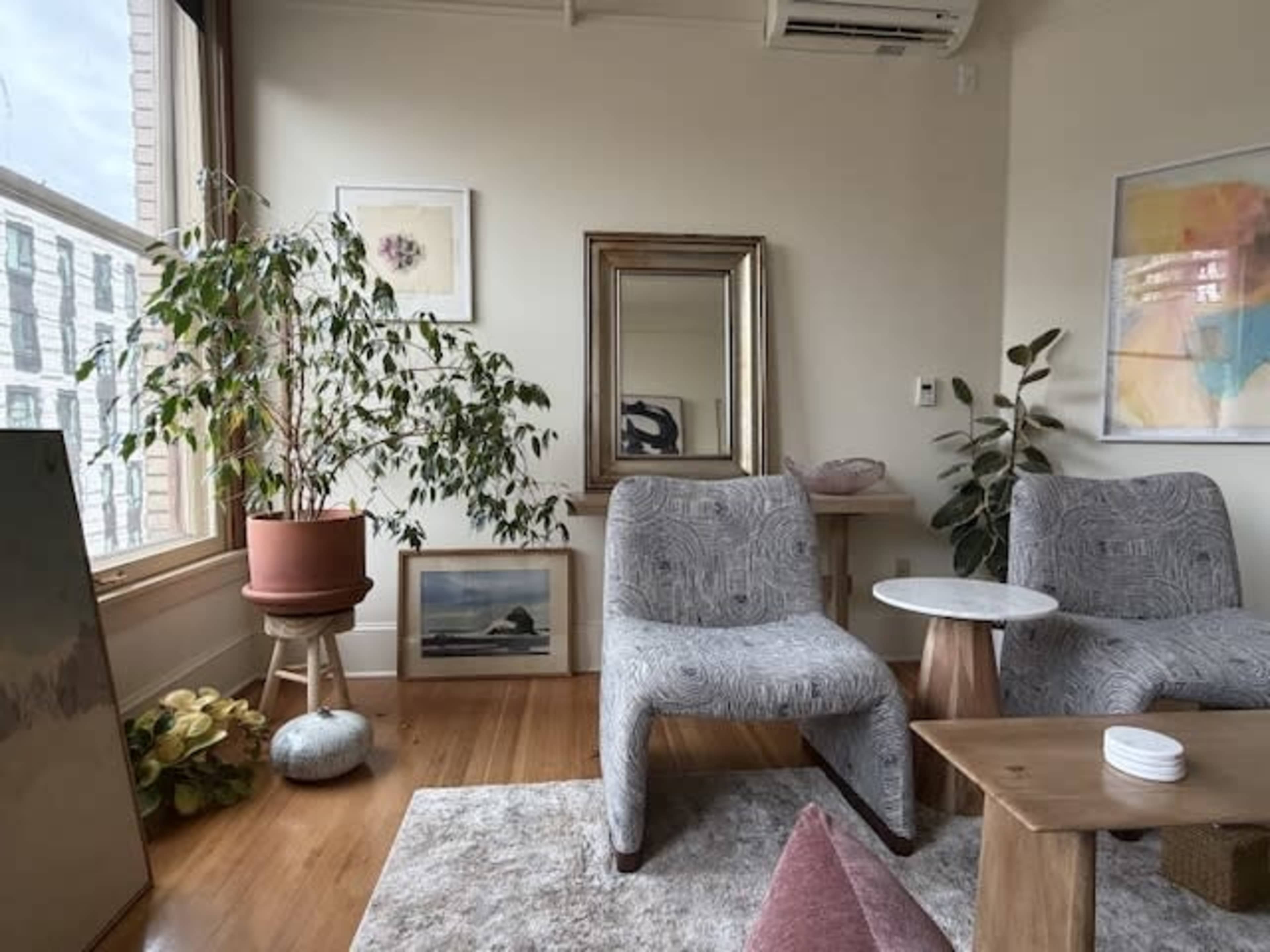 The image shows a cozy room with two patterned chairs, a small side table, a potted plant, artwork on the walls, and a mirror.