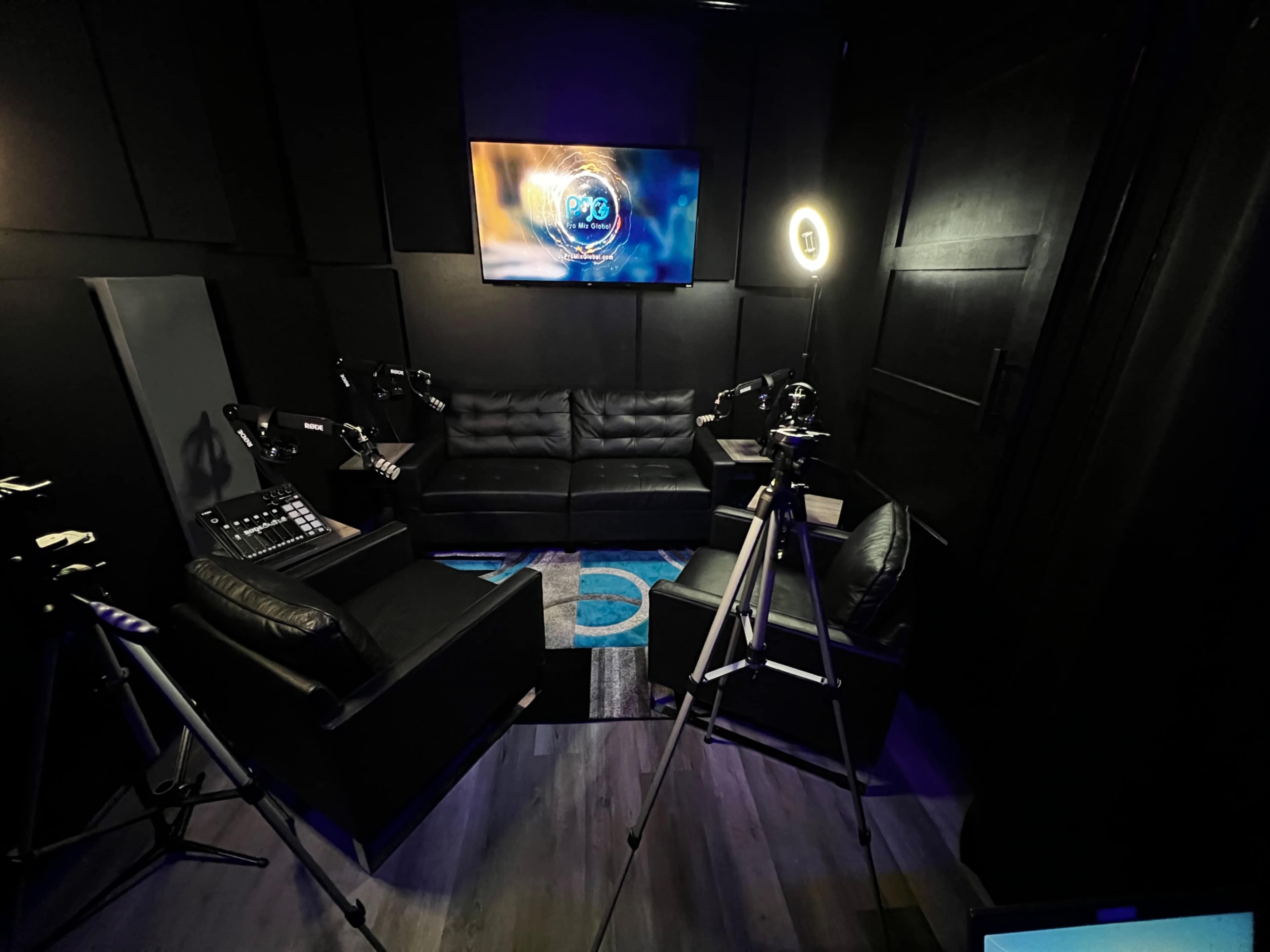 A darkly lit recording studio featuring a couch, several cameras on tripods, and a monitor displaying graphics on the wall.