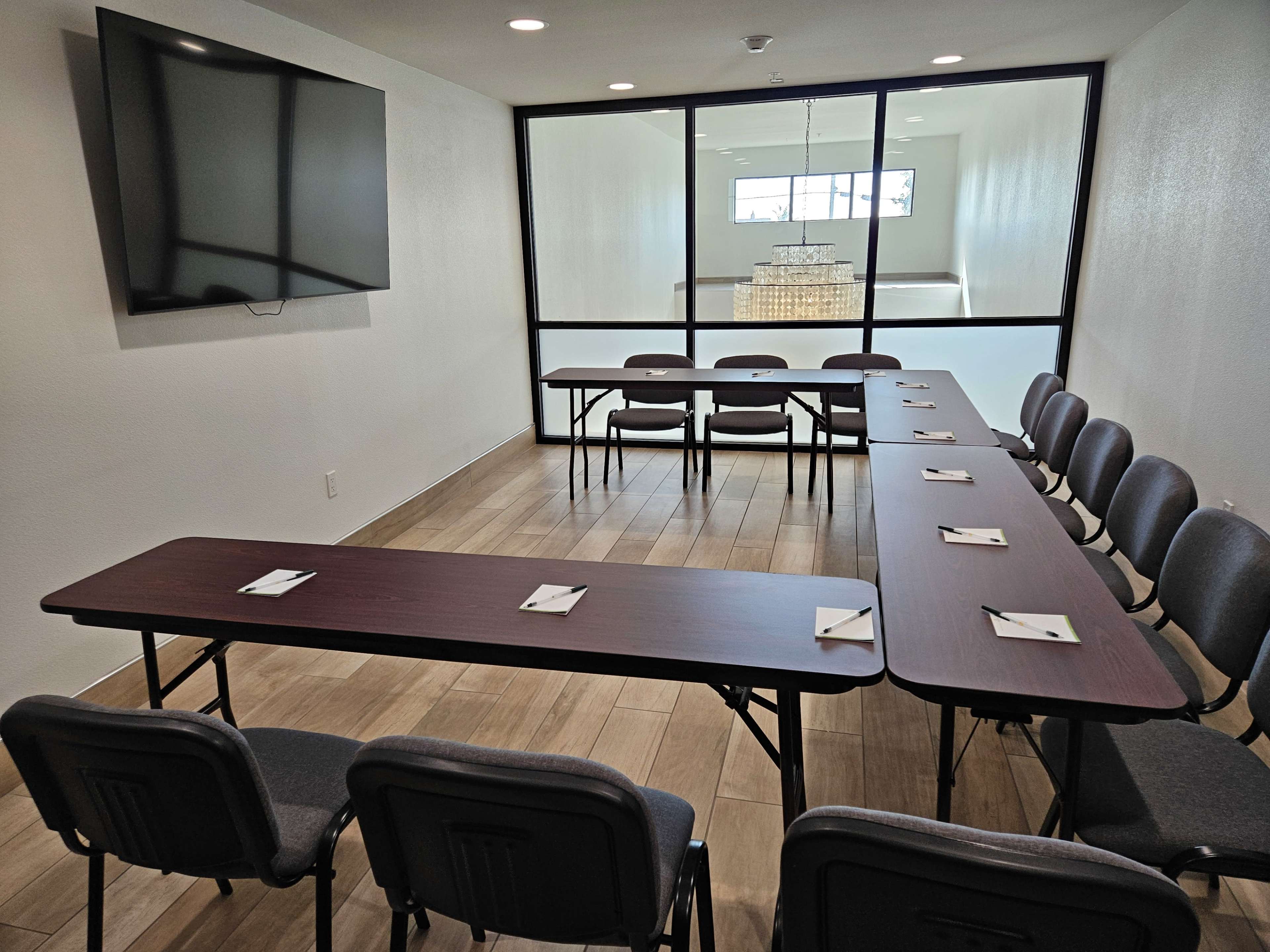 A conference room is arranged with multiple tables and chairs, featuring a wall-mounted TV and a large window.