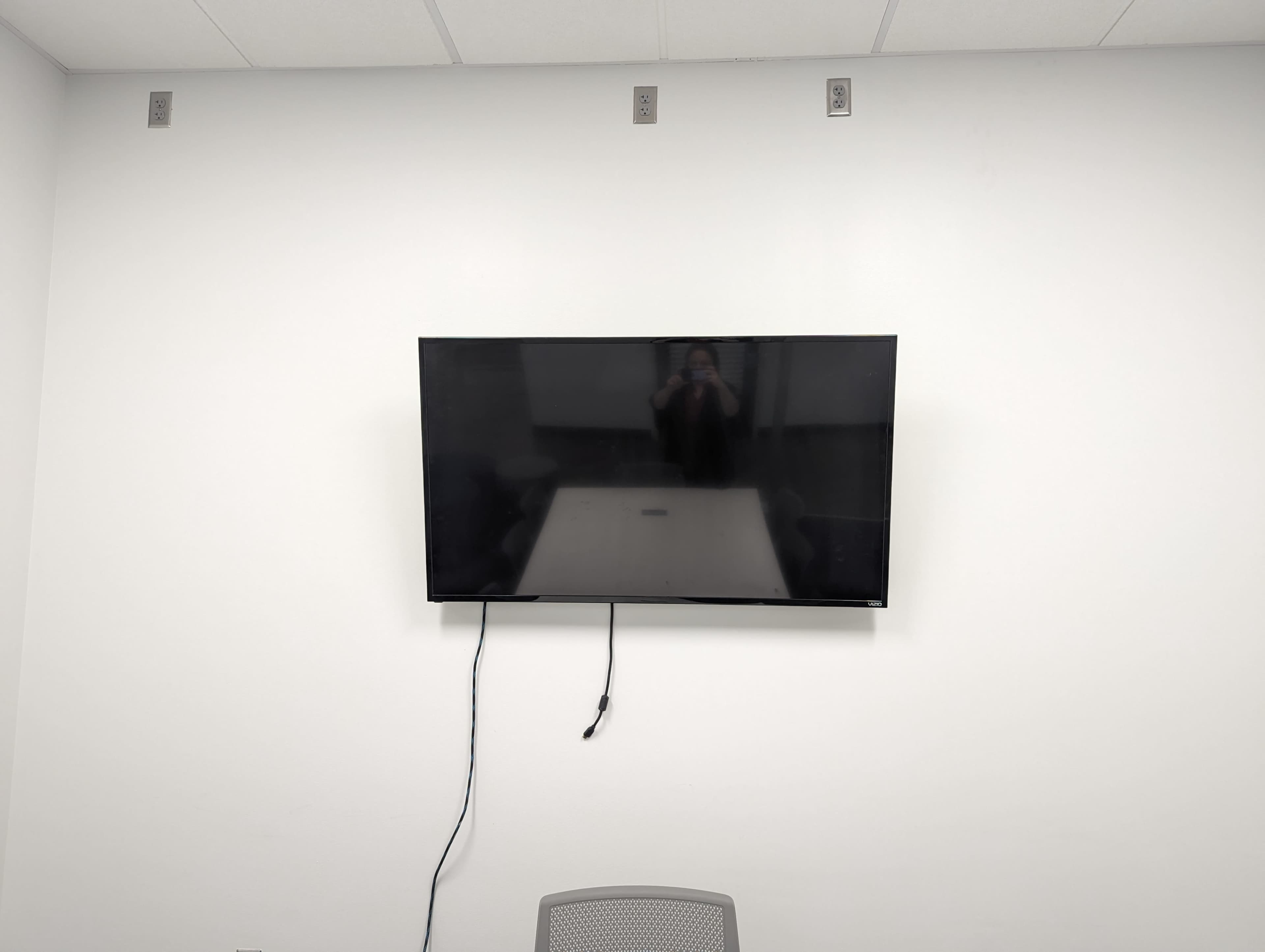 A flat-screen television is mounted on a white wall above a table in a room with a single chair.