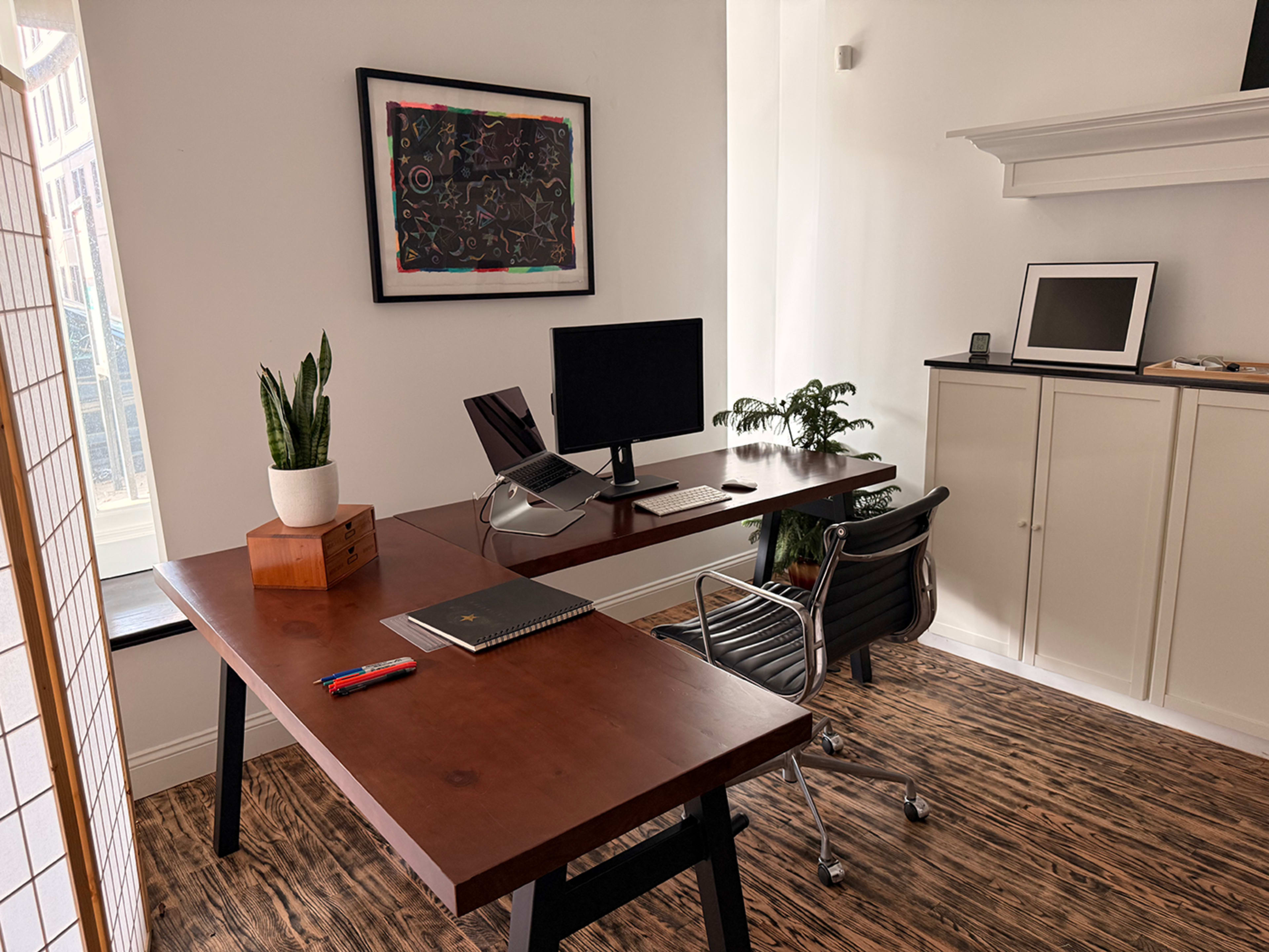 The image shows a modern home office setup with a wooden desk, a computer monitor, stationery, and plants in a bright room.