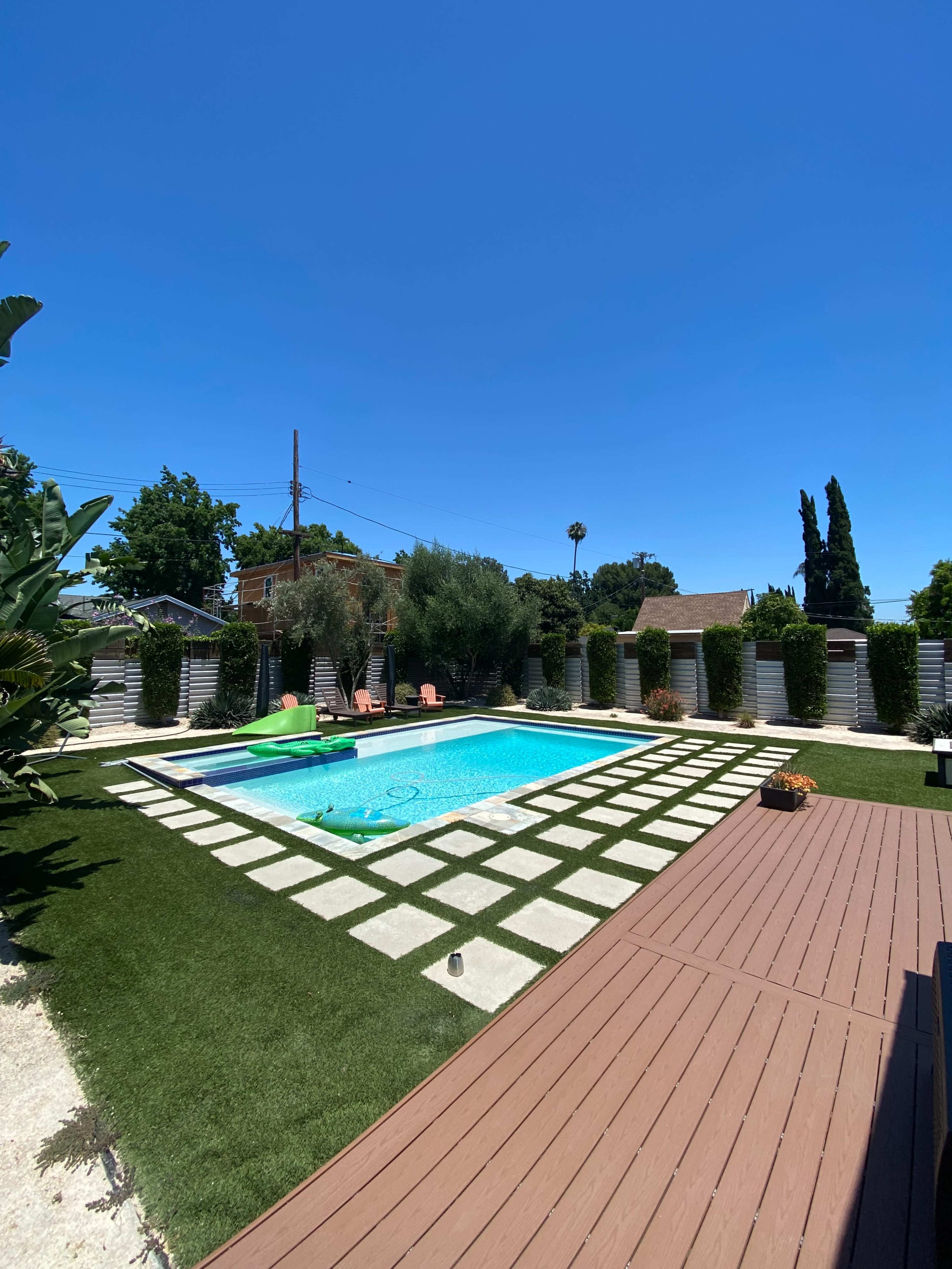 The image shows a backyard with a rectangular swimming pool surrounded by landscaped greenery and a rectangular patio with wooden decking.