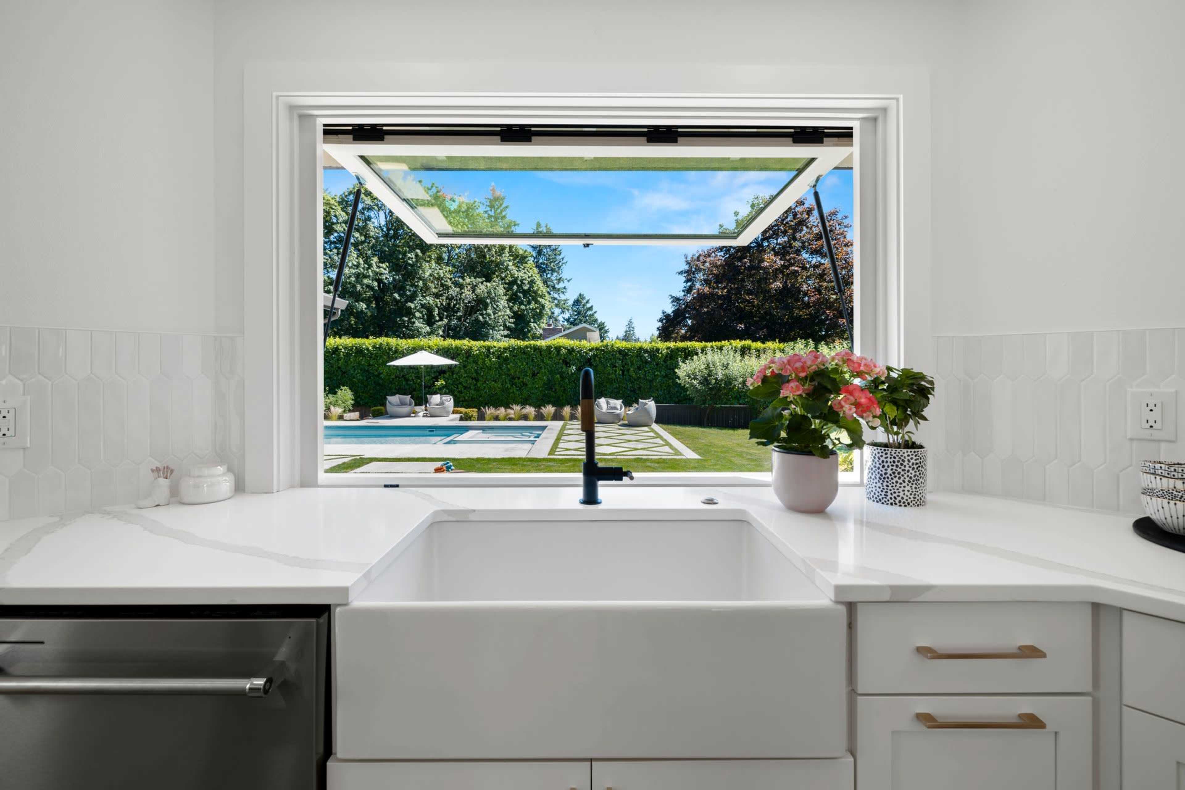 A modern kitchen features a view of a landscaped outdoor pool area through a large open window.