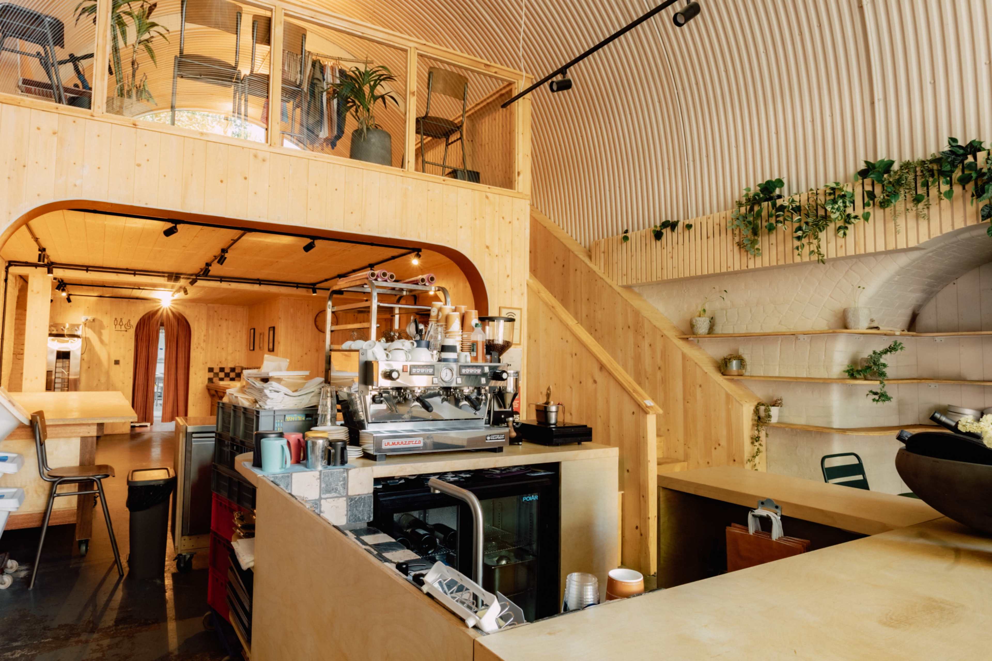 The image shows a modern café interior with wooden walls, a staircase, and a coffee counter featuring an espresso machine.