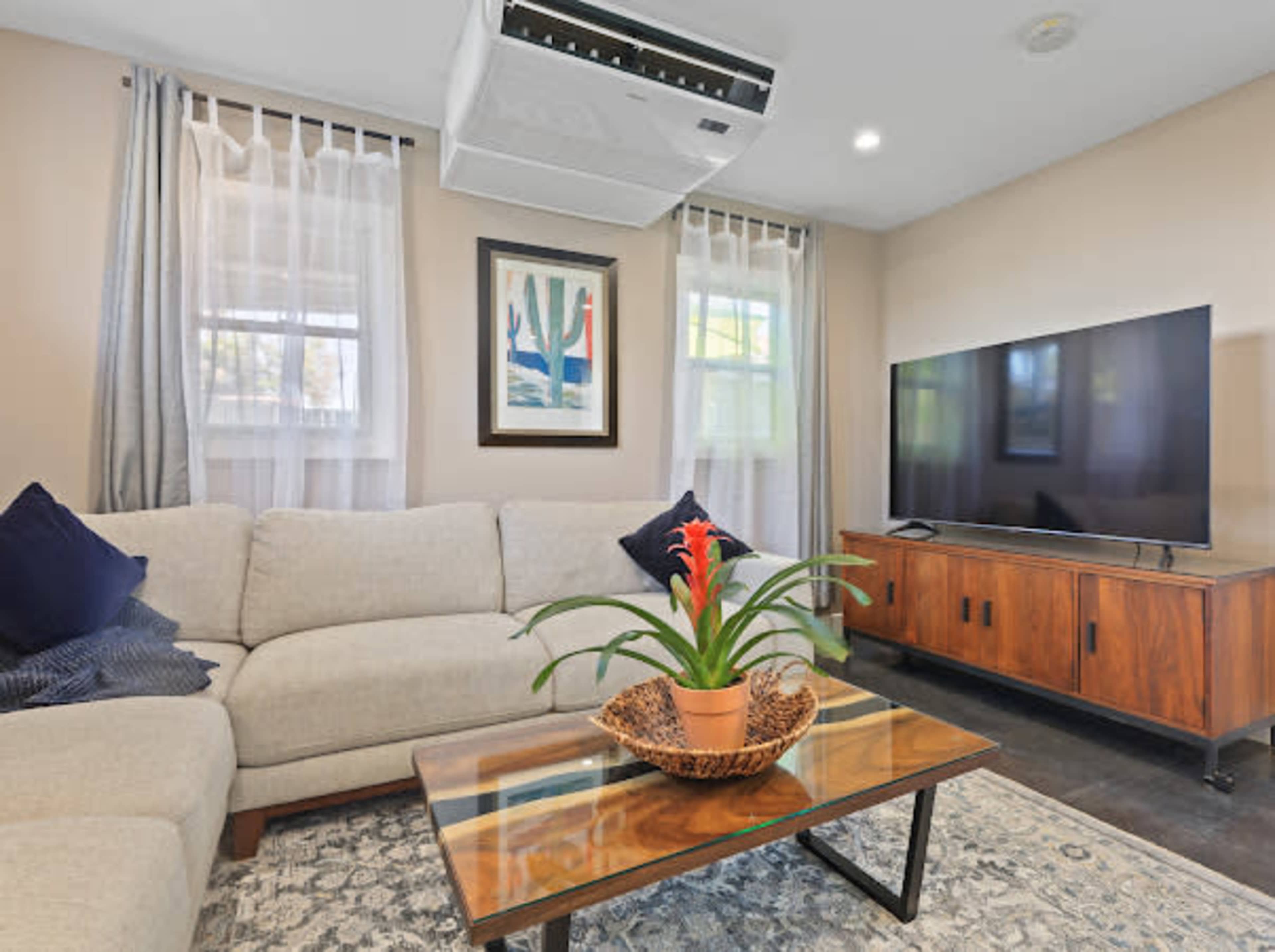 The image shows a modern living room with a sectional couch, a glass coffee table with a plant, a television on a wooden cabinet, and an air conditioning unit mounted on the wall.