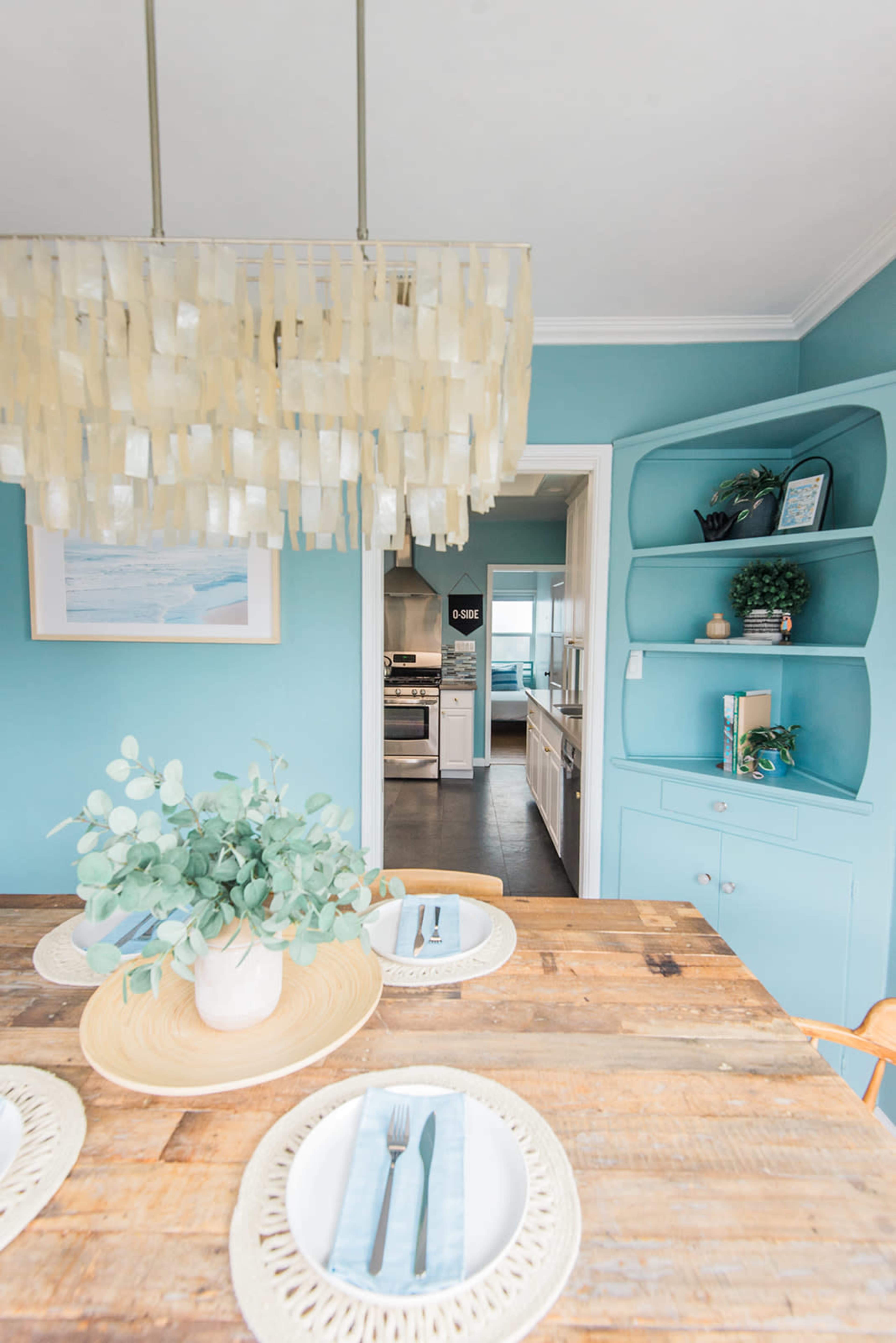 A wooden dining table is set with plates and cutlery, surrounded by chairs, and a light fixture hangs above, while a kitchen is visible through an open doorway.