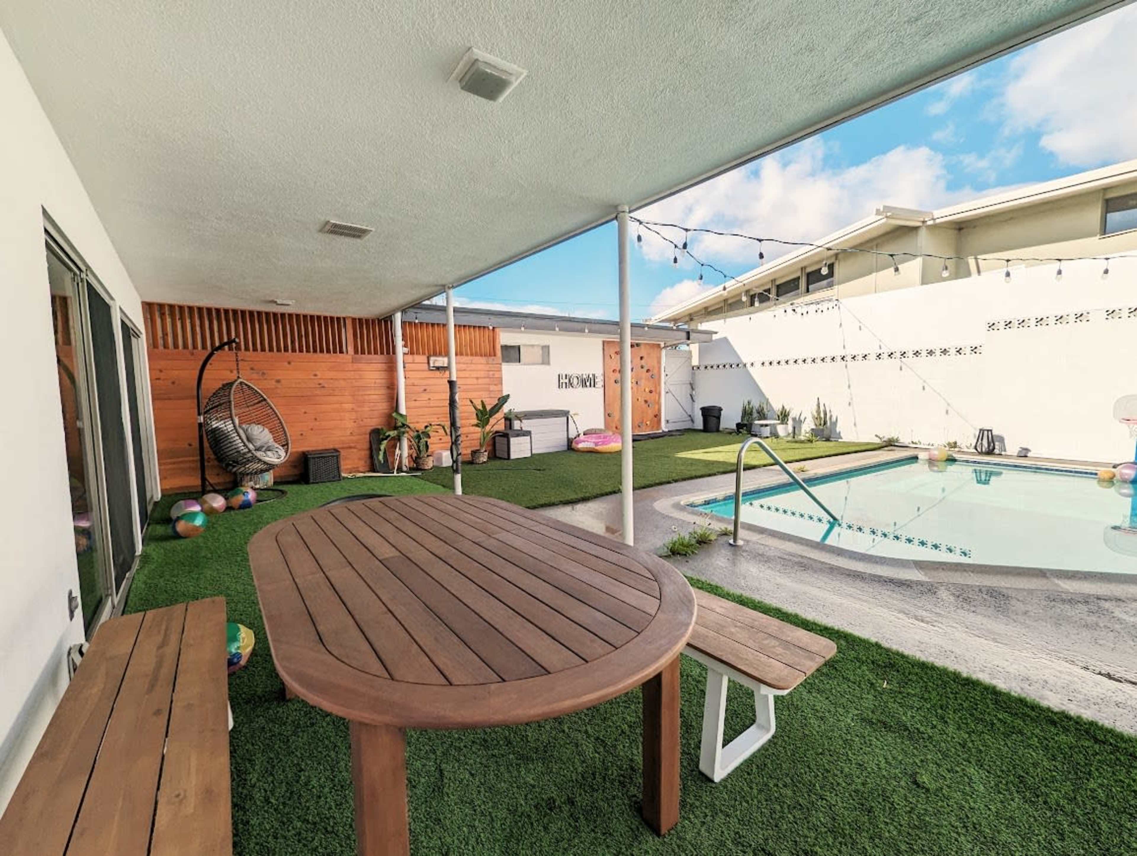 A wooden dining table and bench are set under a covered patio beside a swimming pool surrounded by artificial grass and deck chairs.