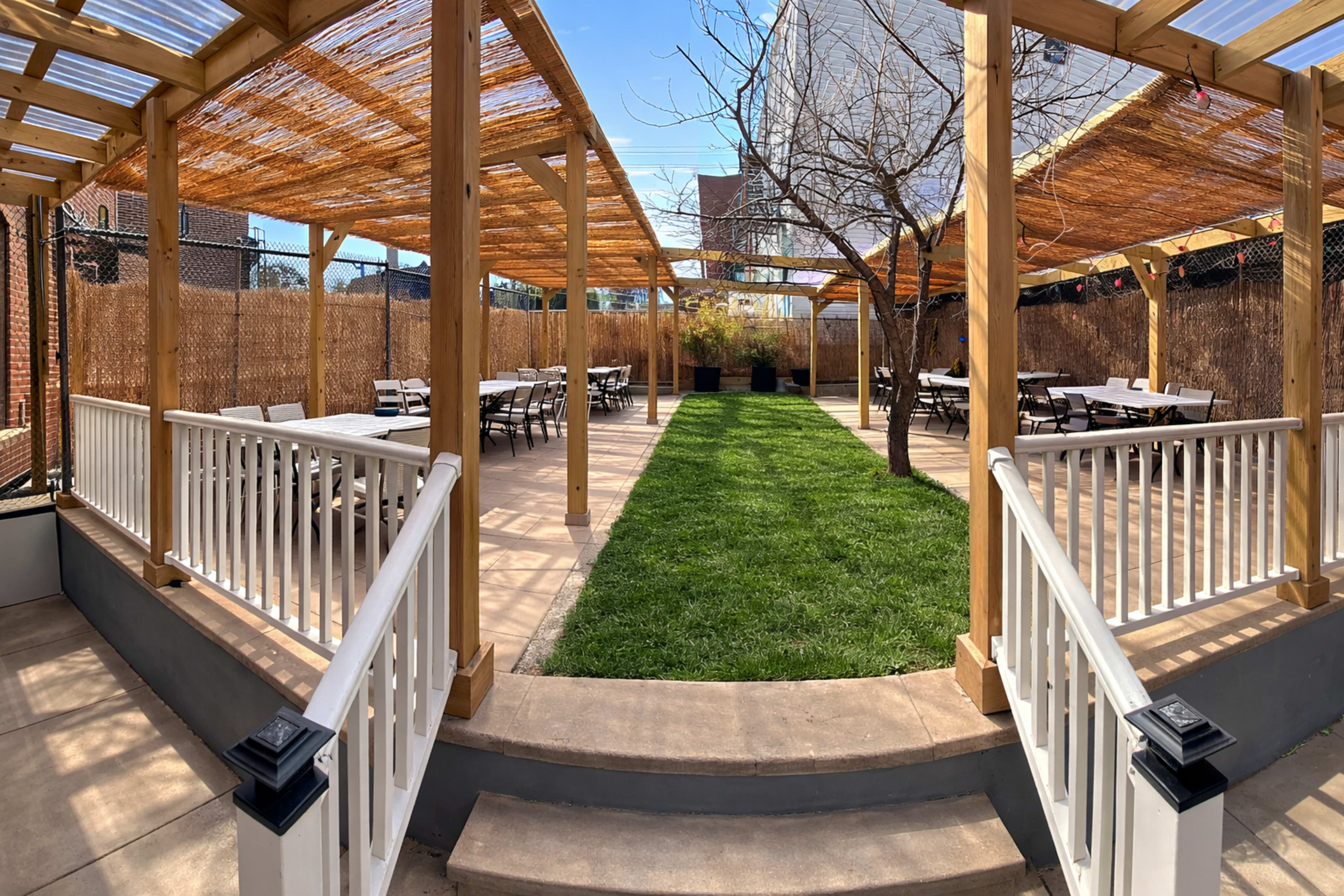 The image shows an outdoor patio area with wooden pergolas, tables arranged for seating, and a grassy central section.