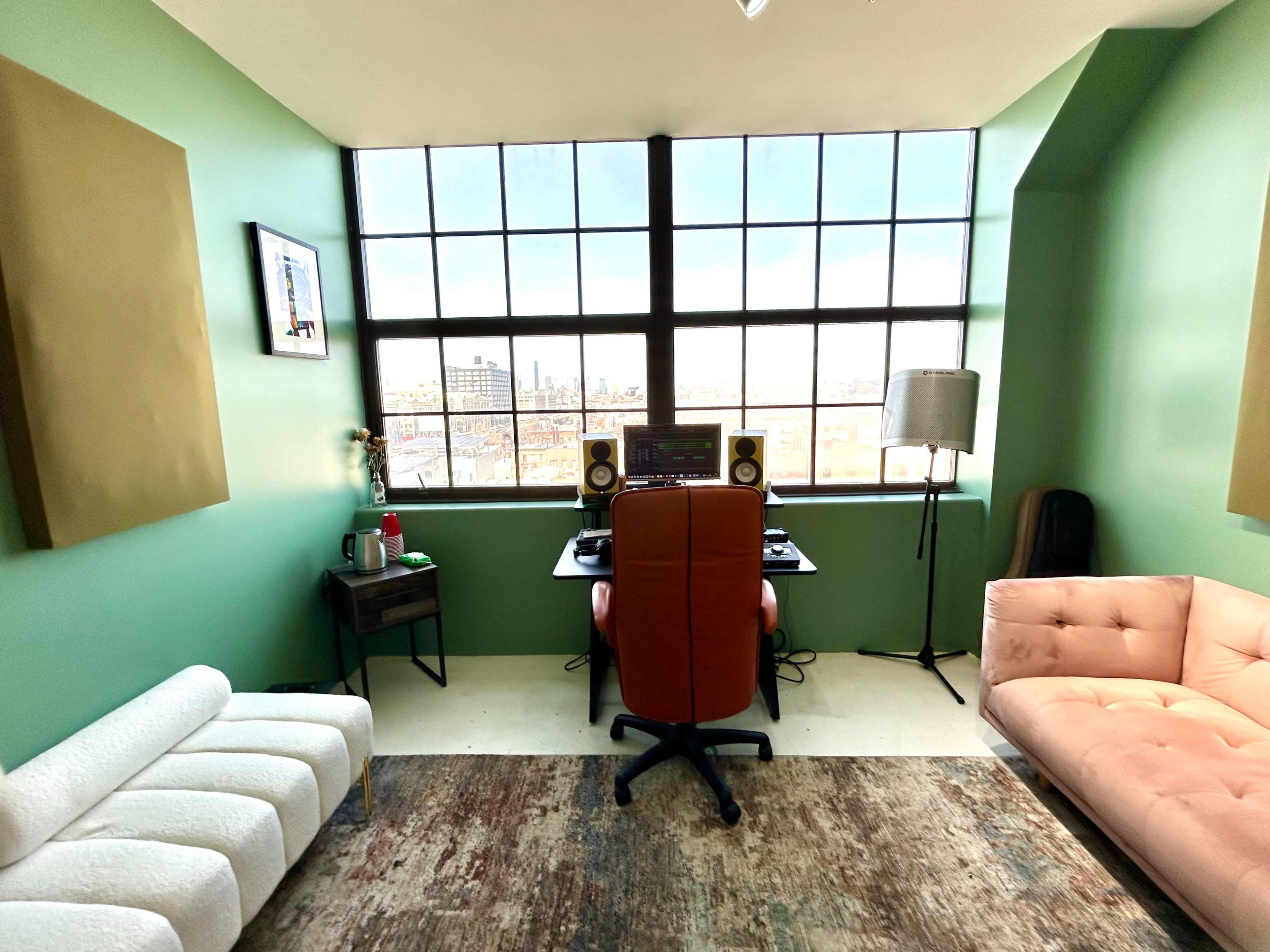The image shows a modern studio space featuring a desk with audio equipment, a brown office chair, and a pink couch, all set against a green wall and a large window overlooking an urban landscape.