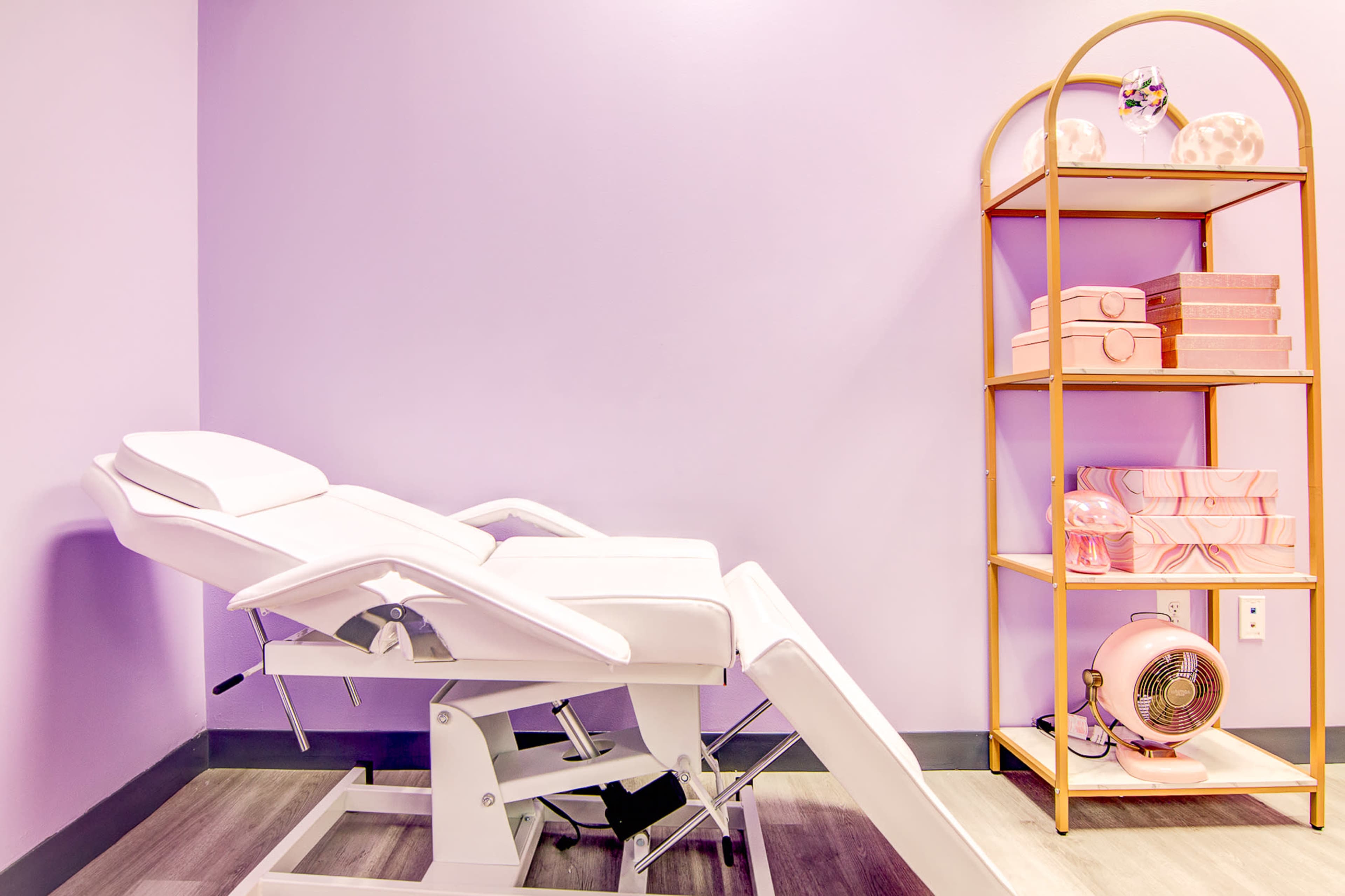 A white treatment chair is positioned beside a gold shelving unit holding pink boxes and a small fan, against a lavender wall.