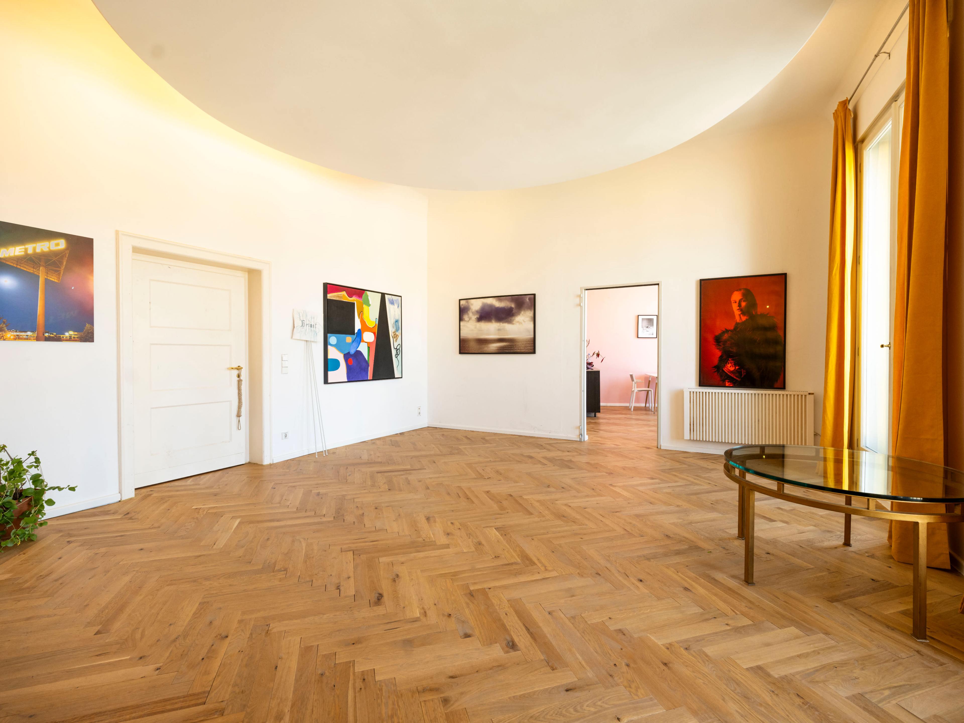 The image shows a spacious, well-lit room with wooden herringbone flooring and several framed artworks on the walls.