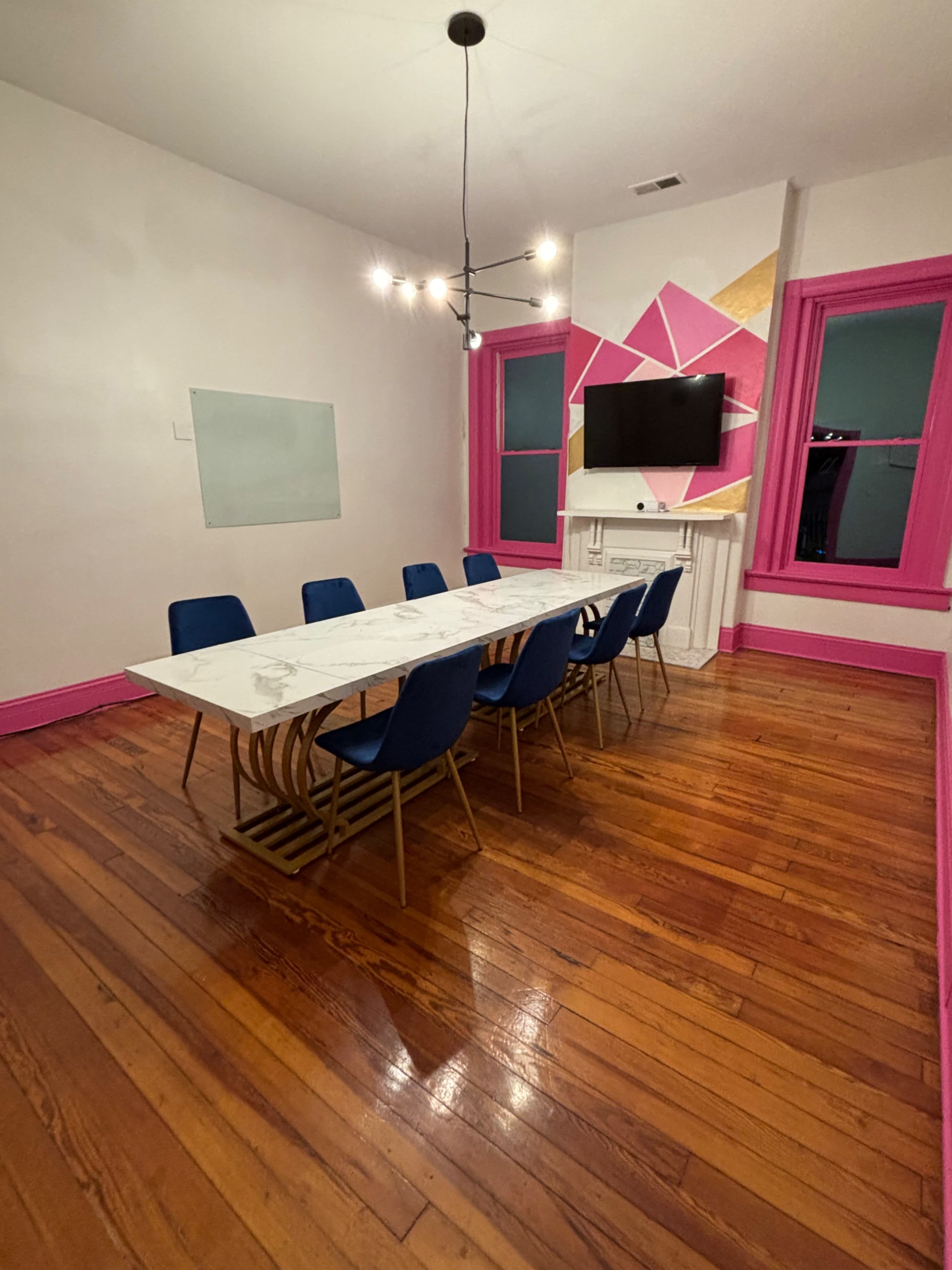 A modern conference room features a marble table surrounded by eight blue chairs, with a television mounted on a pink accent wall.