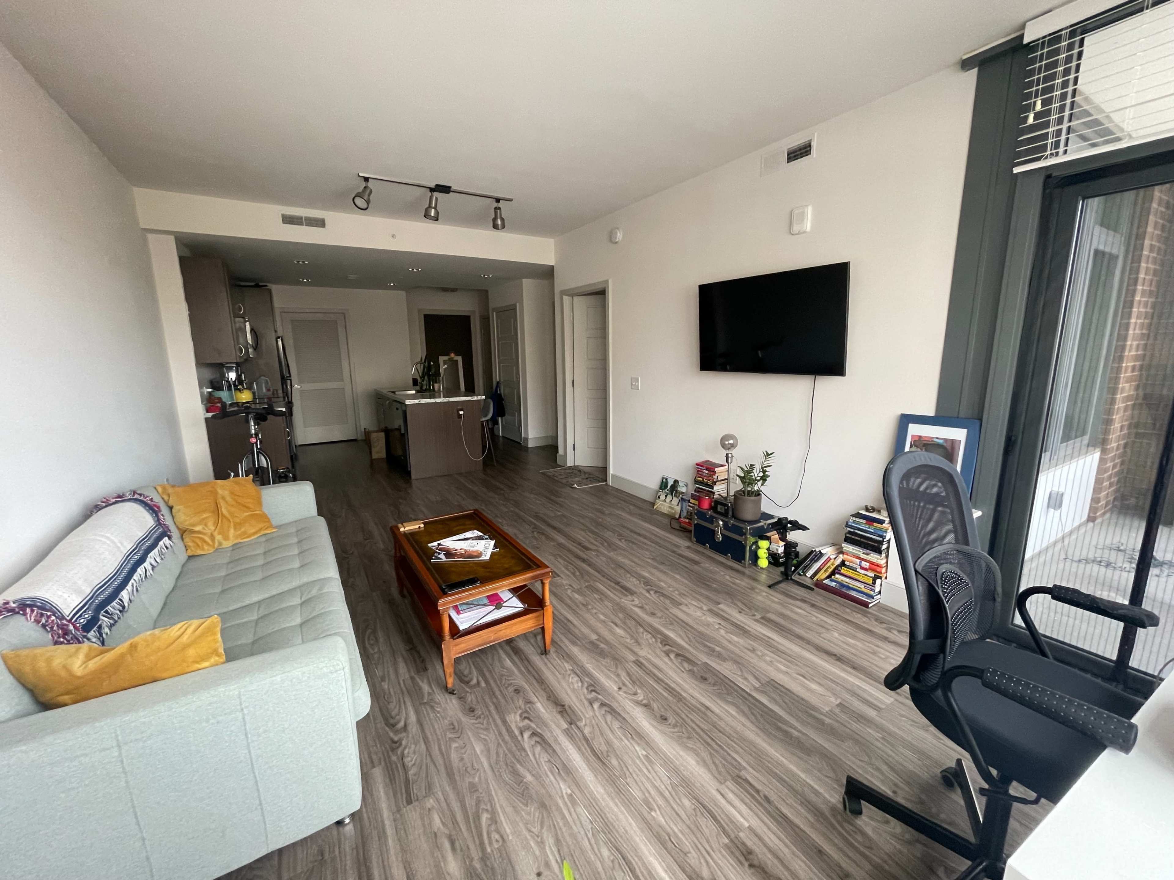 A modern, light-filled apartment features a gray sofa, a wooden coffee table, and a large wall-mounted television, with an open kitchen area visible in the background.