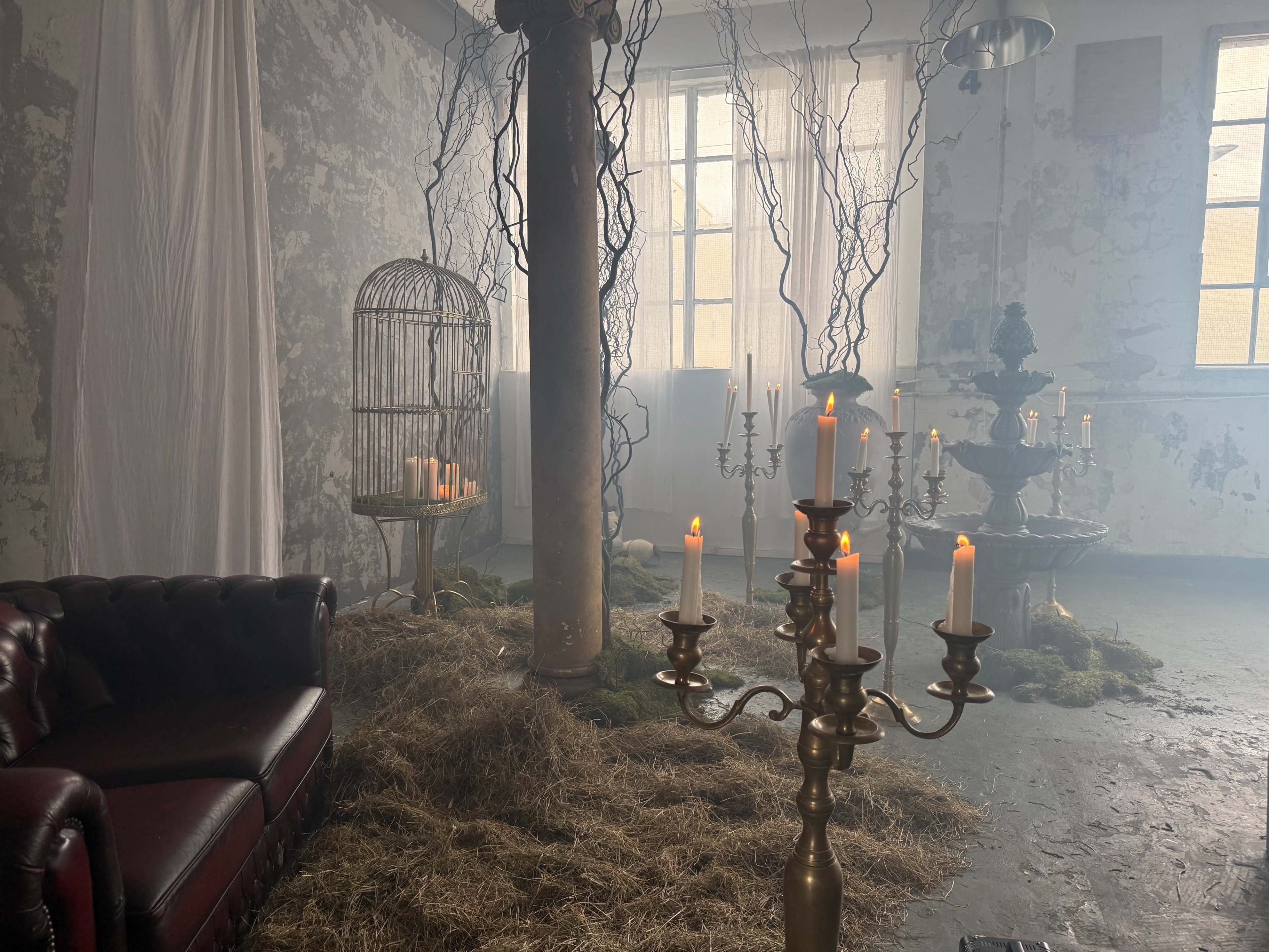 The scene features a dimly lit room with a vintage leather couch, a decorative column, a birdcage, a fountain, and several candles placed among patches of straw and moss on the floor.