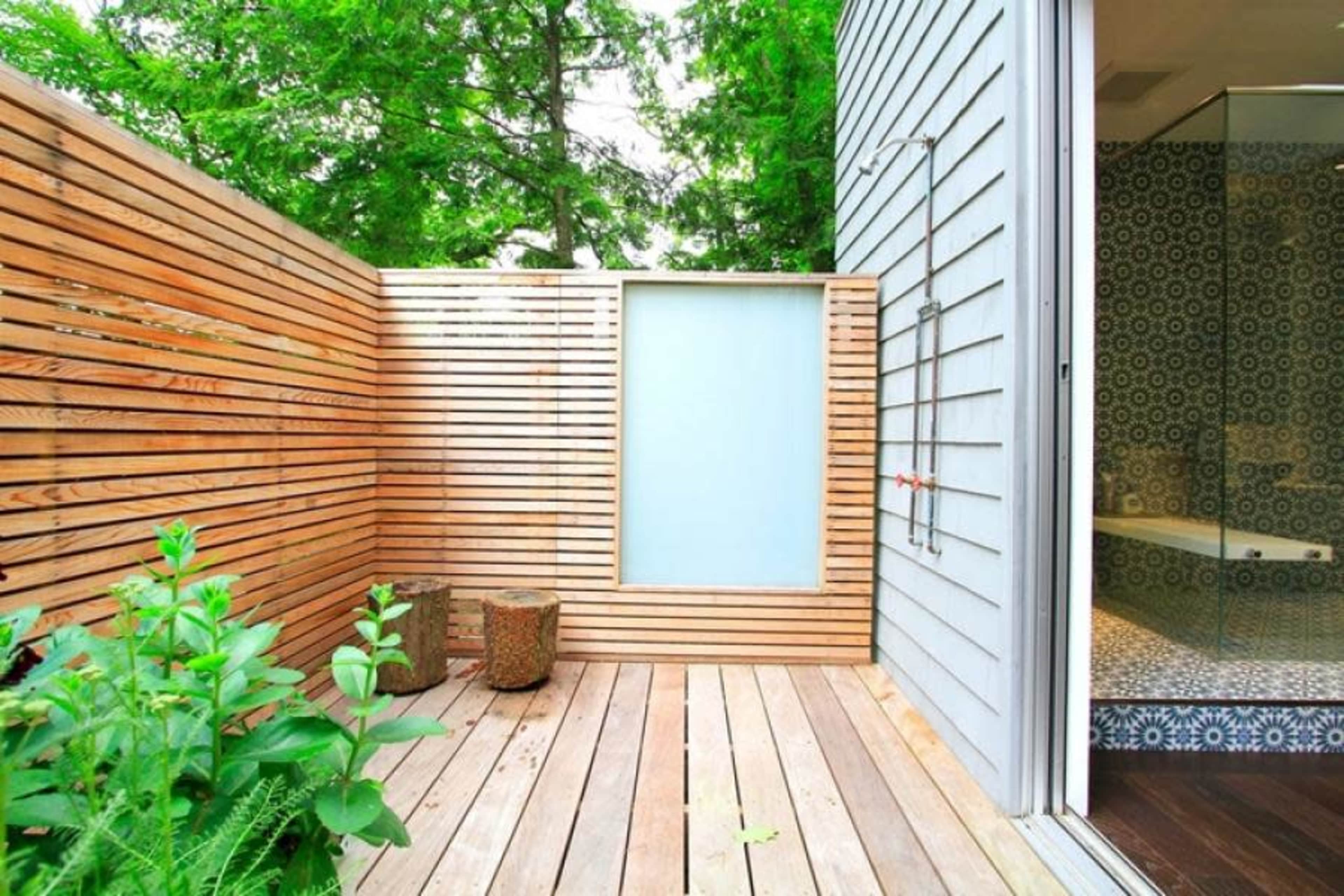 The image shows a wooden terrace enclosed by slatted fencing, with a glass panel, potted plants, and a view of an adjoining bathroom area.