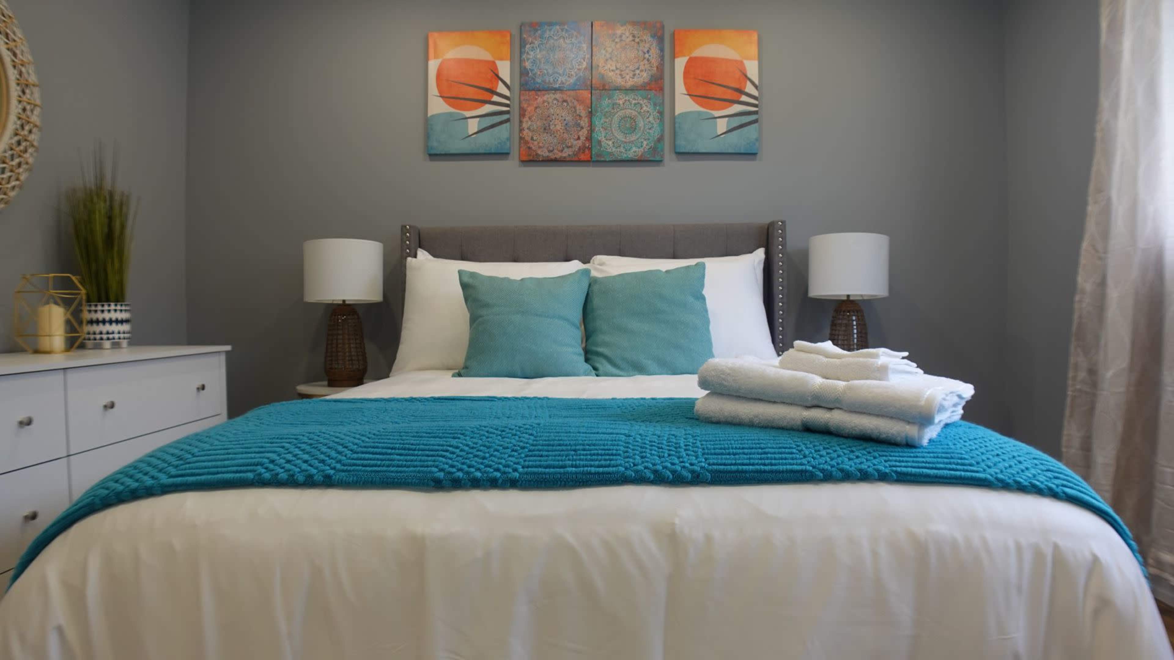 A neatly made bed with turquoise accents and folded towels is positioned in a well-decorated bedroom featuring artwork on the wall.