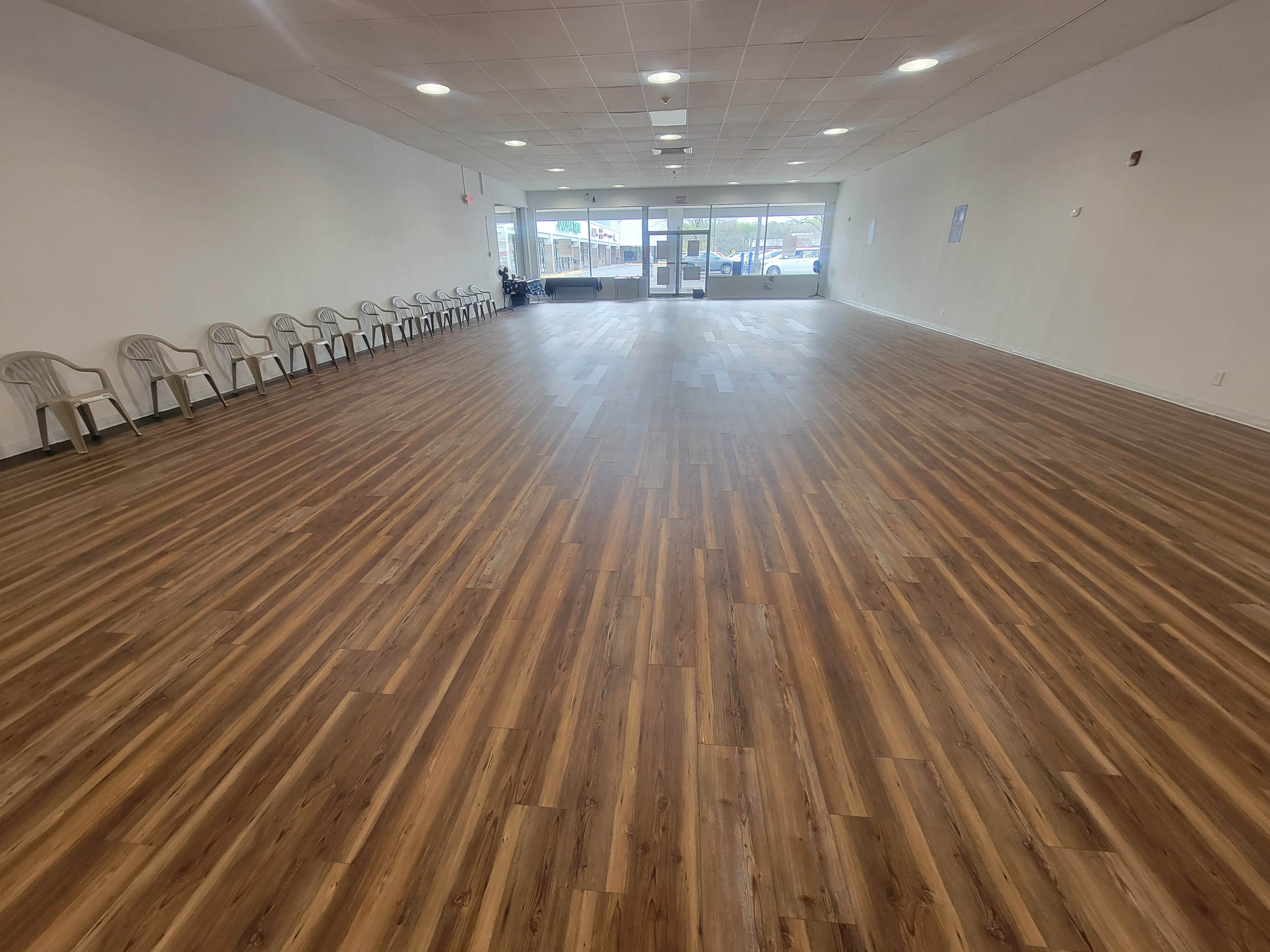 An empty room with a polished wooden floor and a row of plastic chairs along one wall, featuring large windows at the entrance.