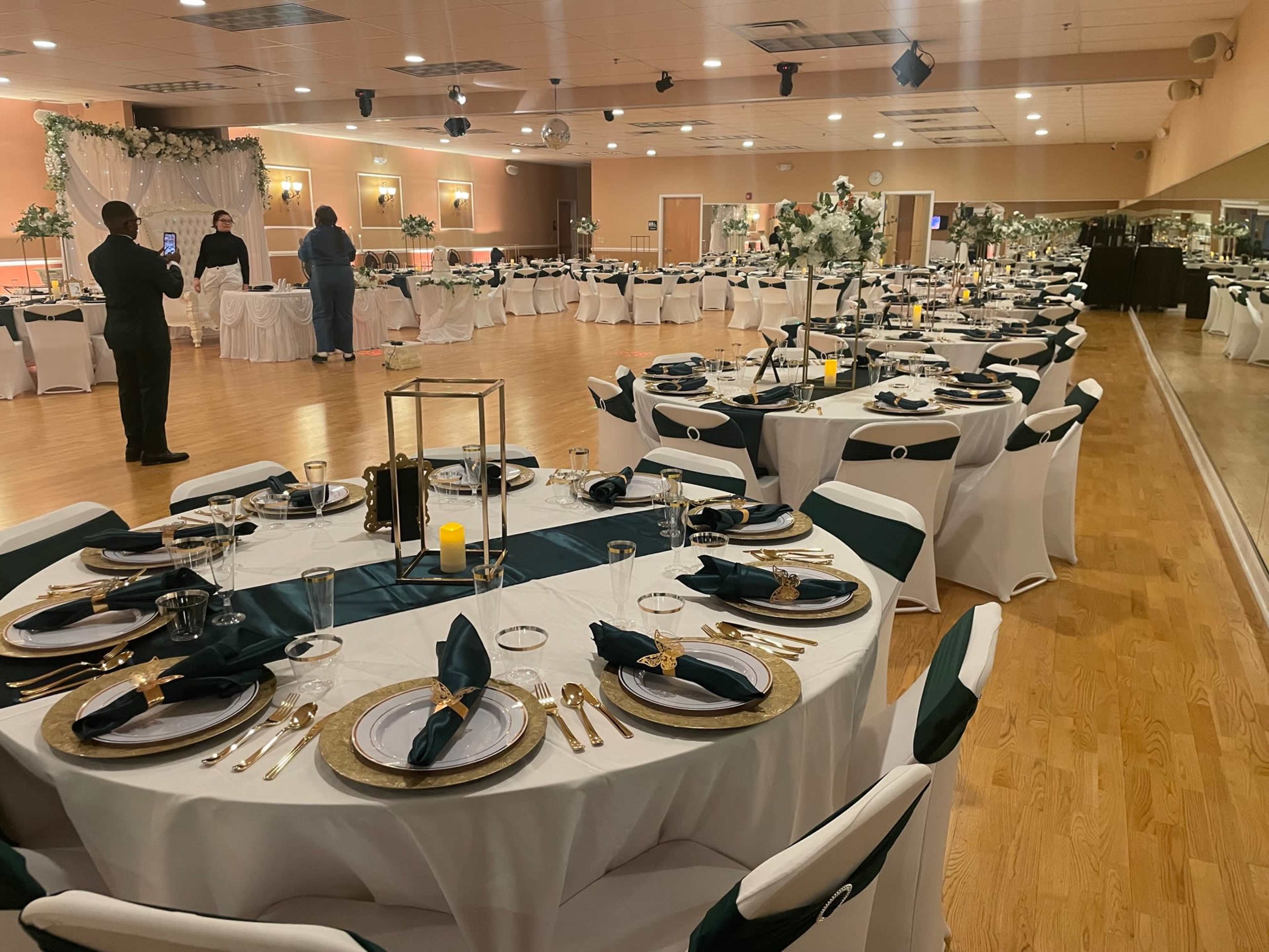 The image shows a banquet hall set up for an event, with multiple elegantly arranged tables, each covered in white tablecloths and decorated with gold and green accents.