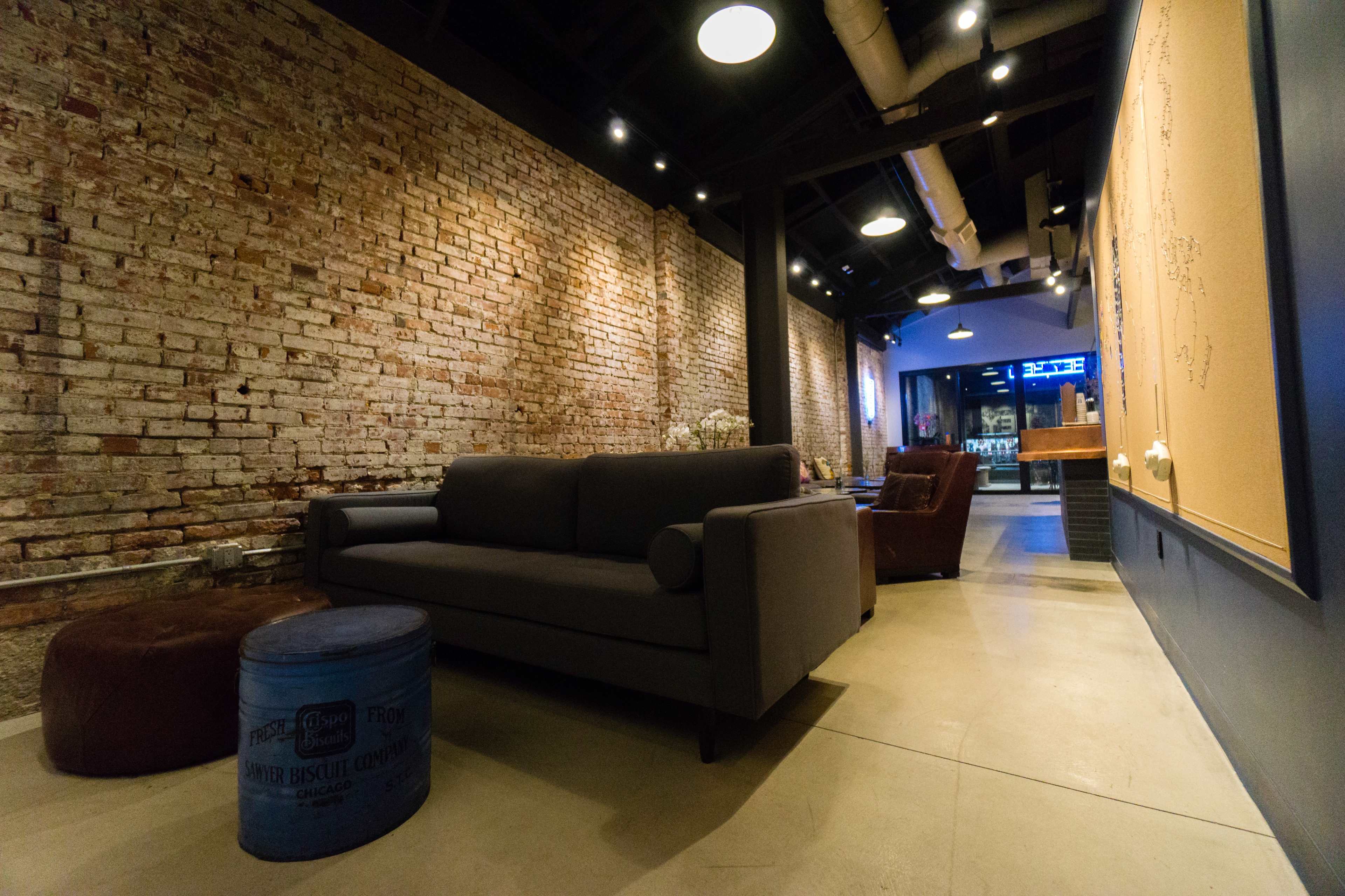 A modern interior features a gray sofa, a brown armchair, and a blue vintage trunk against a backdrop of exposed brick walls and concrete flooring.