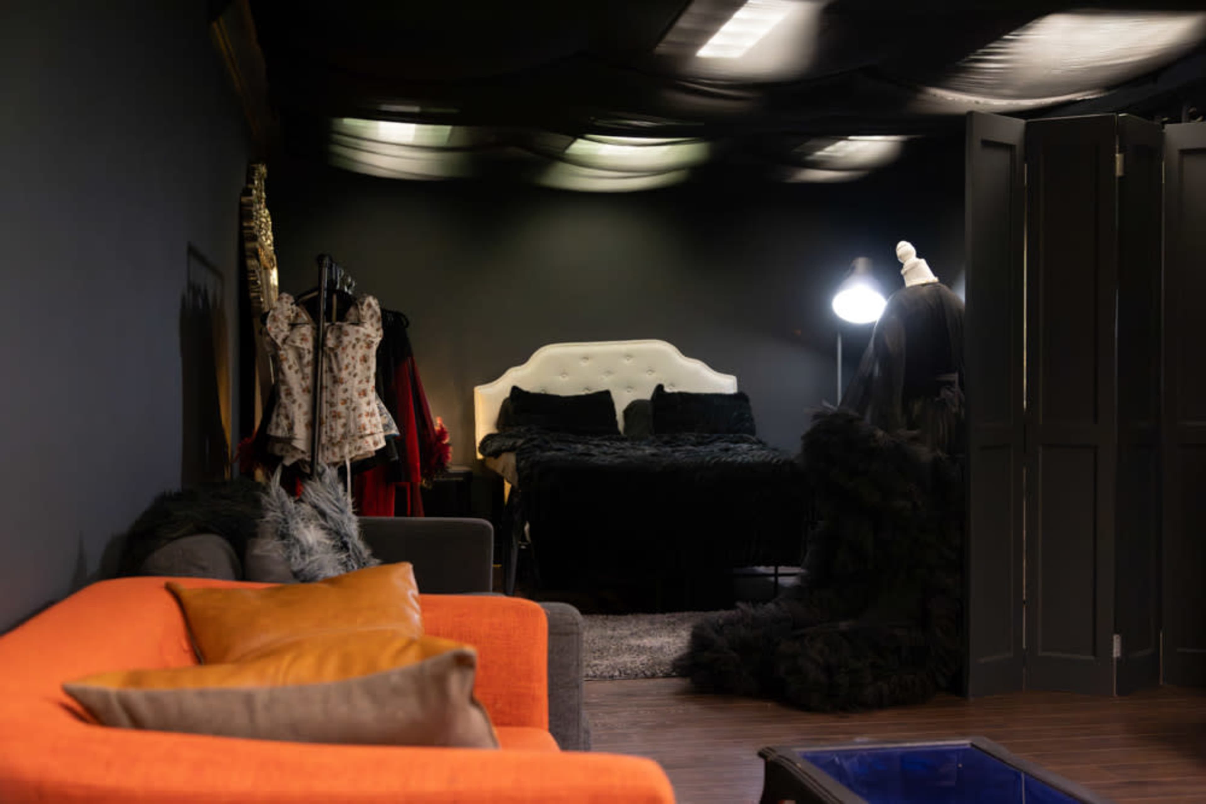 A dimly lit room featuring a black wall, a cozy orange couch with pillows, a bed with dark bedding, and several clothing racks displaying various garments.