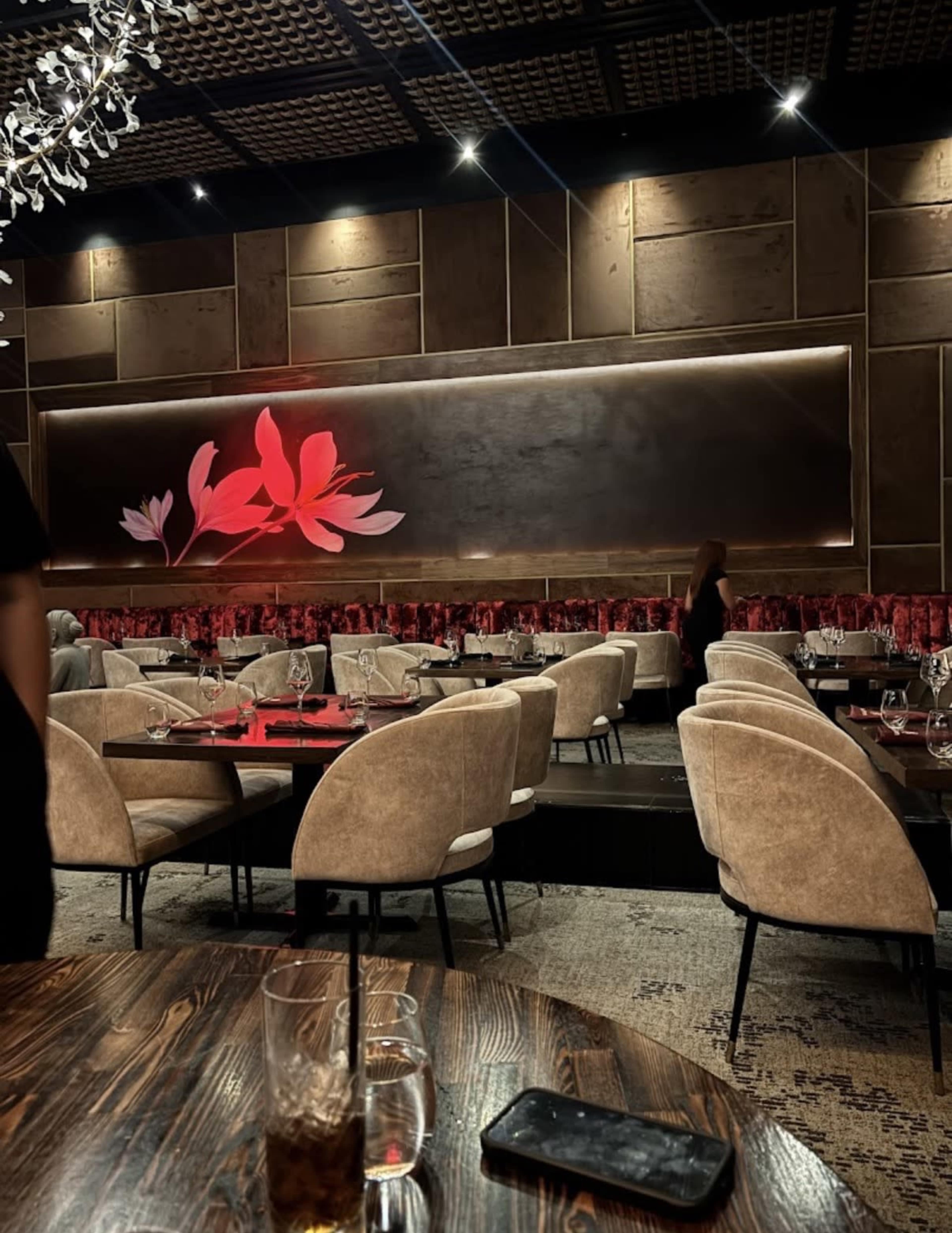 The image shows a dimly lit restaurant interior with beige upholstered chairs, wooden tables, and a large floral artwork on the wall.