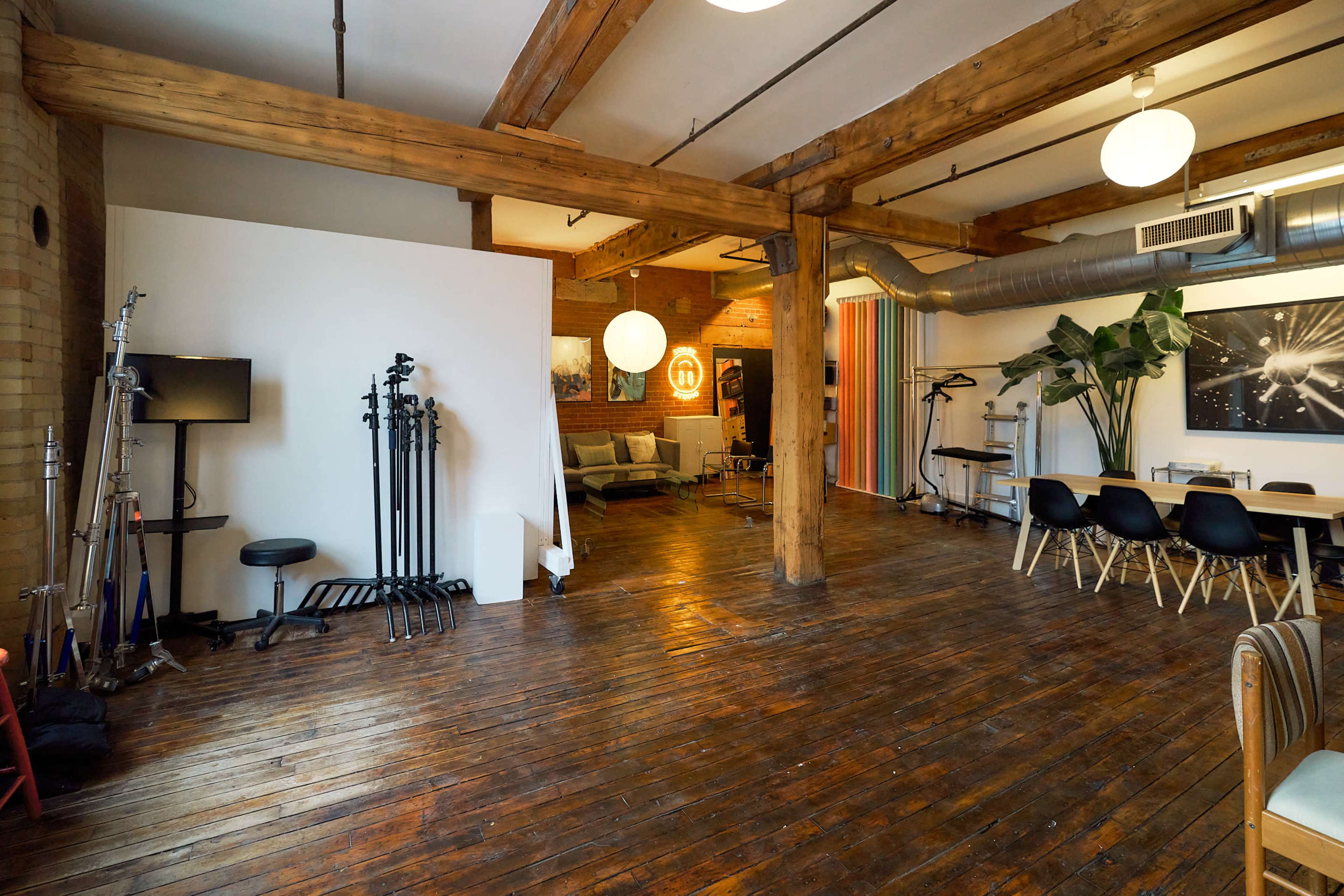 The image shows a spacious interior with wooden beams, a polished wooden floor, and various standing equipment, including lighting stands, alongside a seating area and a dining table.