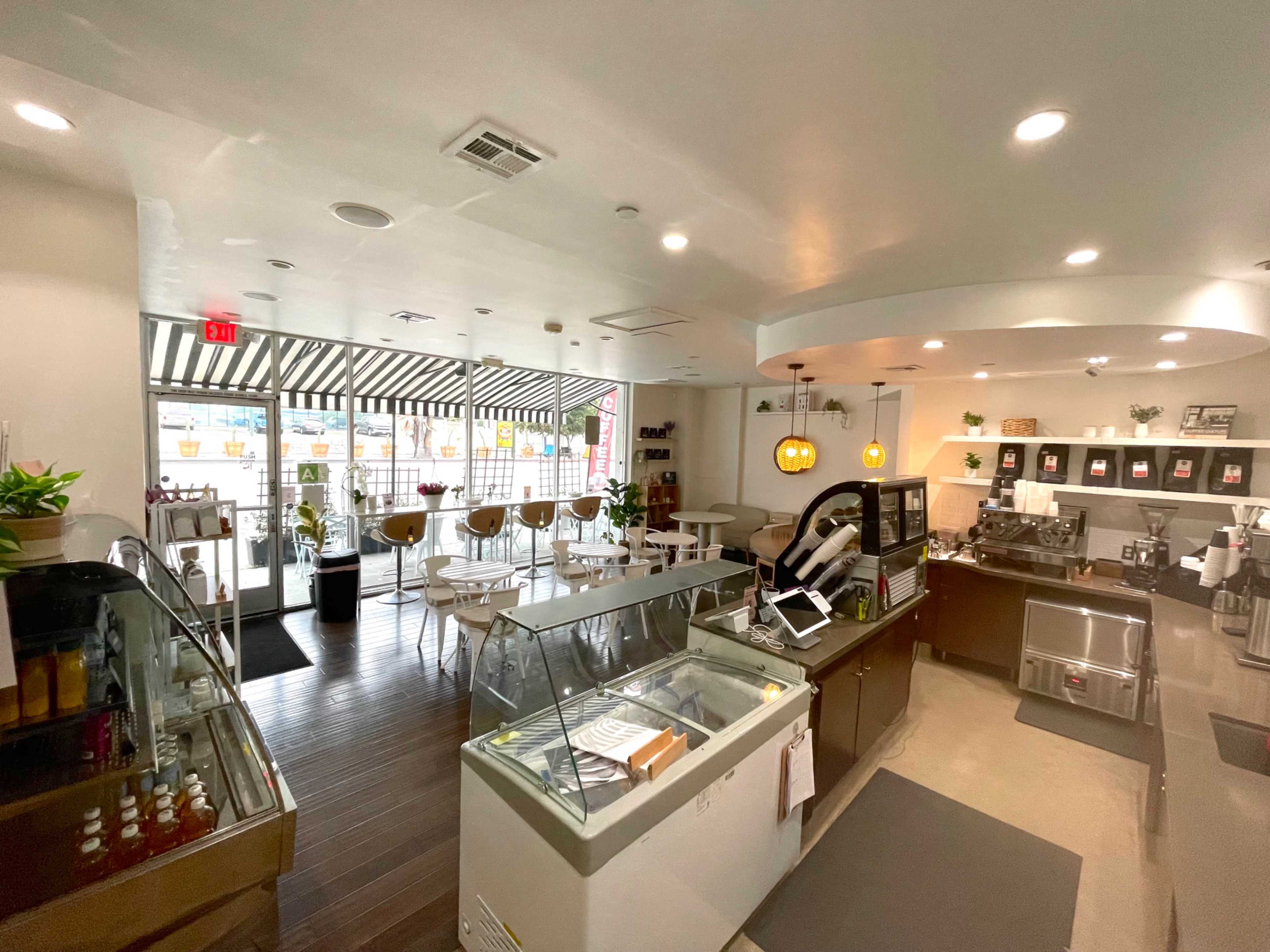 The image shows the interior of a modern café with seating, a display case for desserts, and large windows letting in natural light.