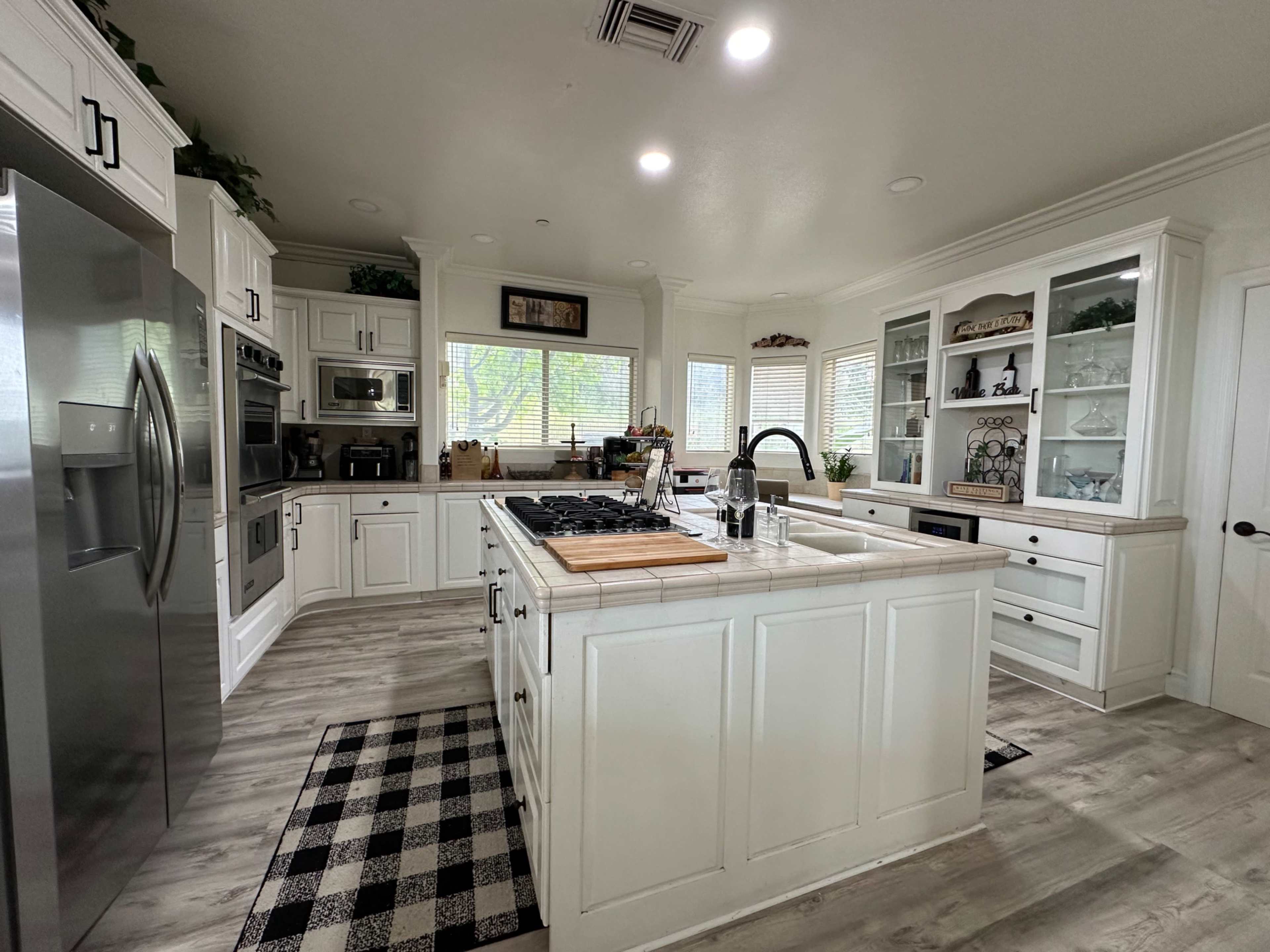 The kitchen features white cabinetry, a central island with a cooktop, and large windows allowing natural light to enter the space.