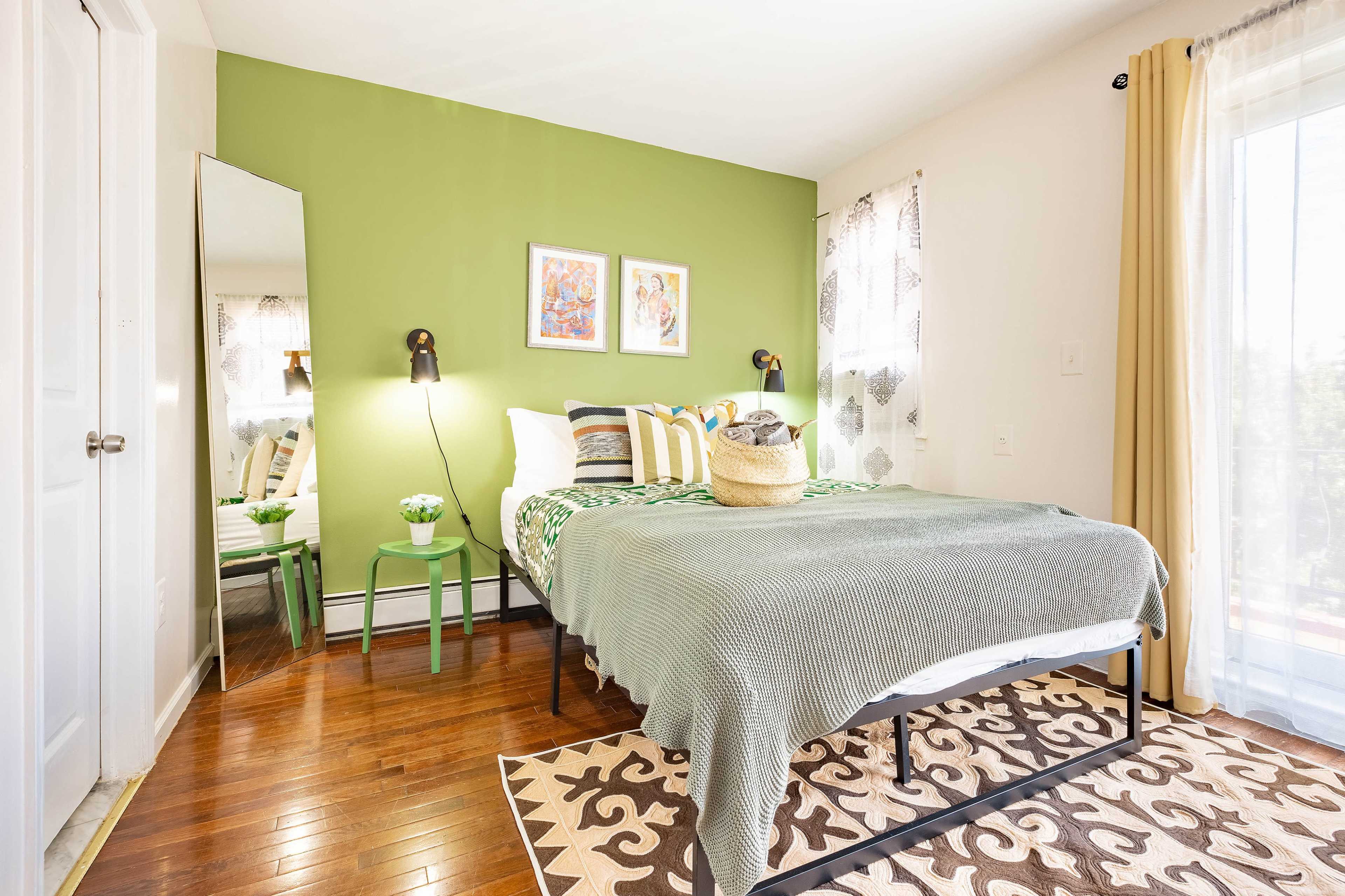 The image shows a bedroom featuring a bed with green and white bedding, a large mirror, two framed artworks on the wall, and patterned curtains.