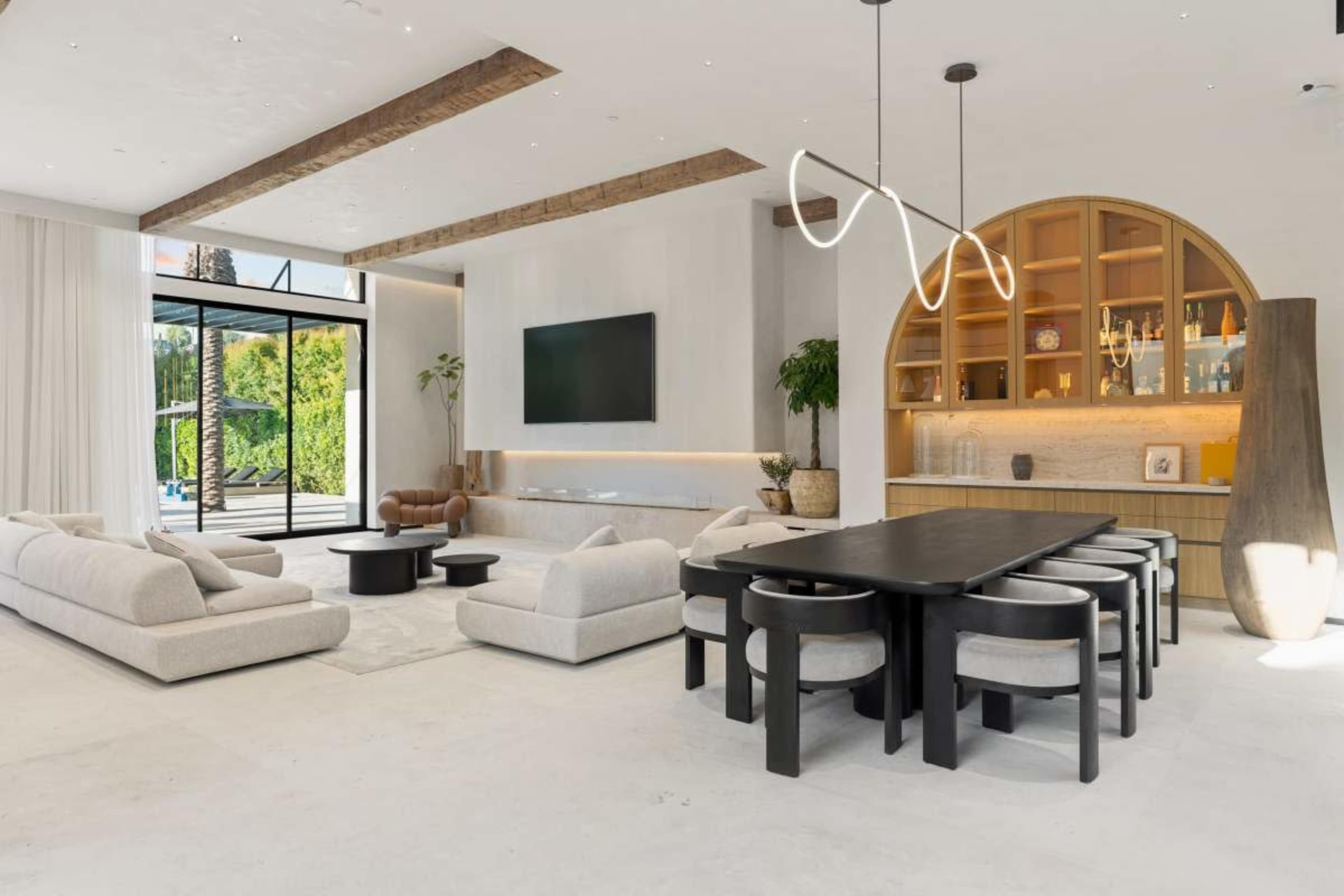 The image shows a modern living space featuring a large dining table, a cozy seating area with sofas, and a decorative wall unit with shelves, all bathed in natural light from large windows.