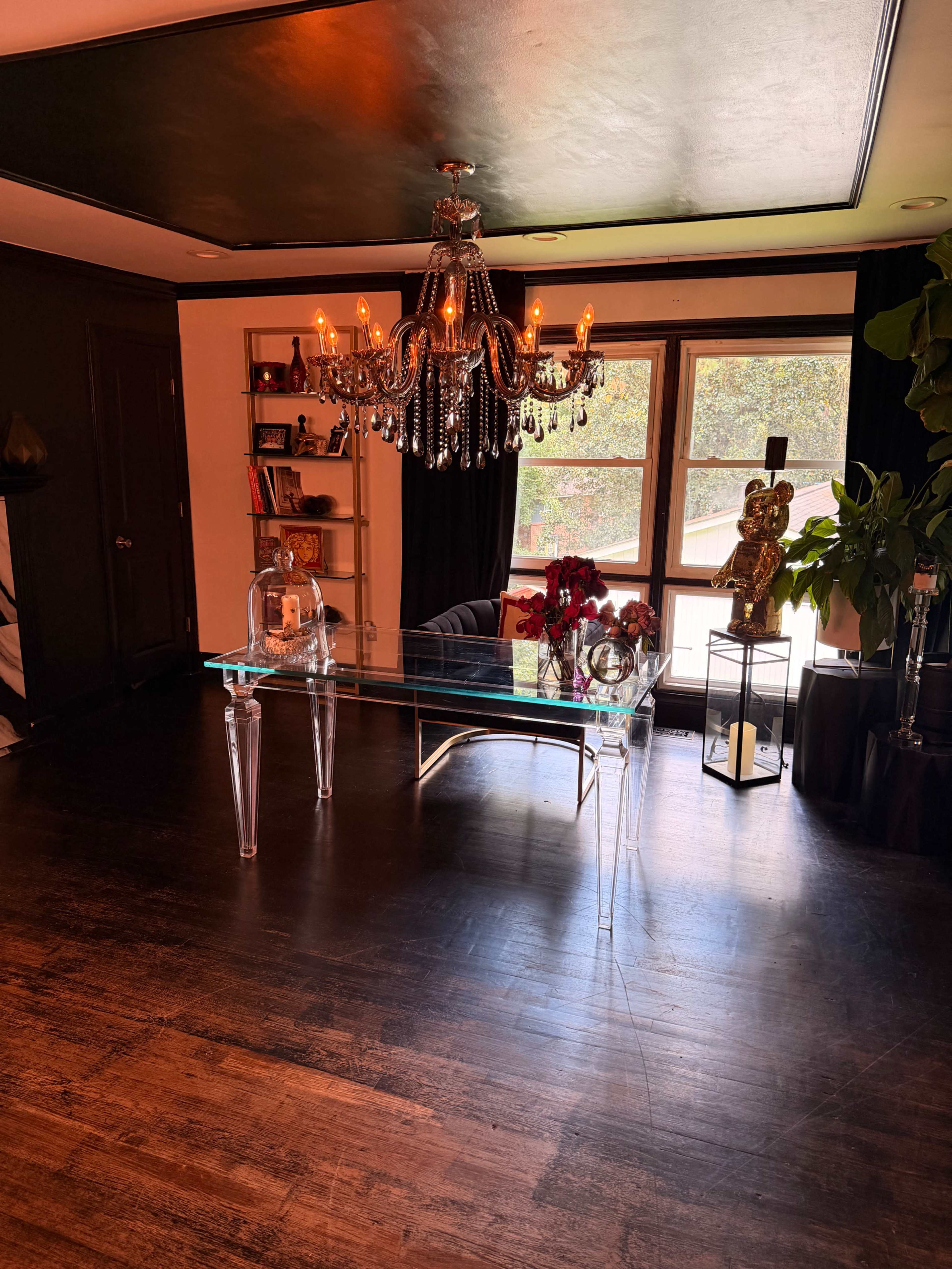 The image shows a modern room featuring a glass desk under a crystal chandelier, with black curtains and potted plants in the background.