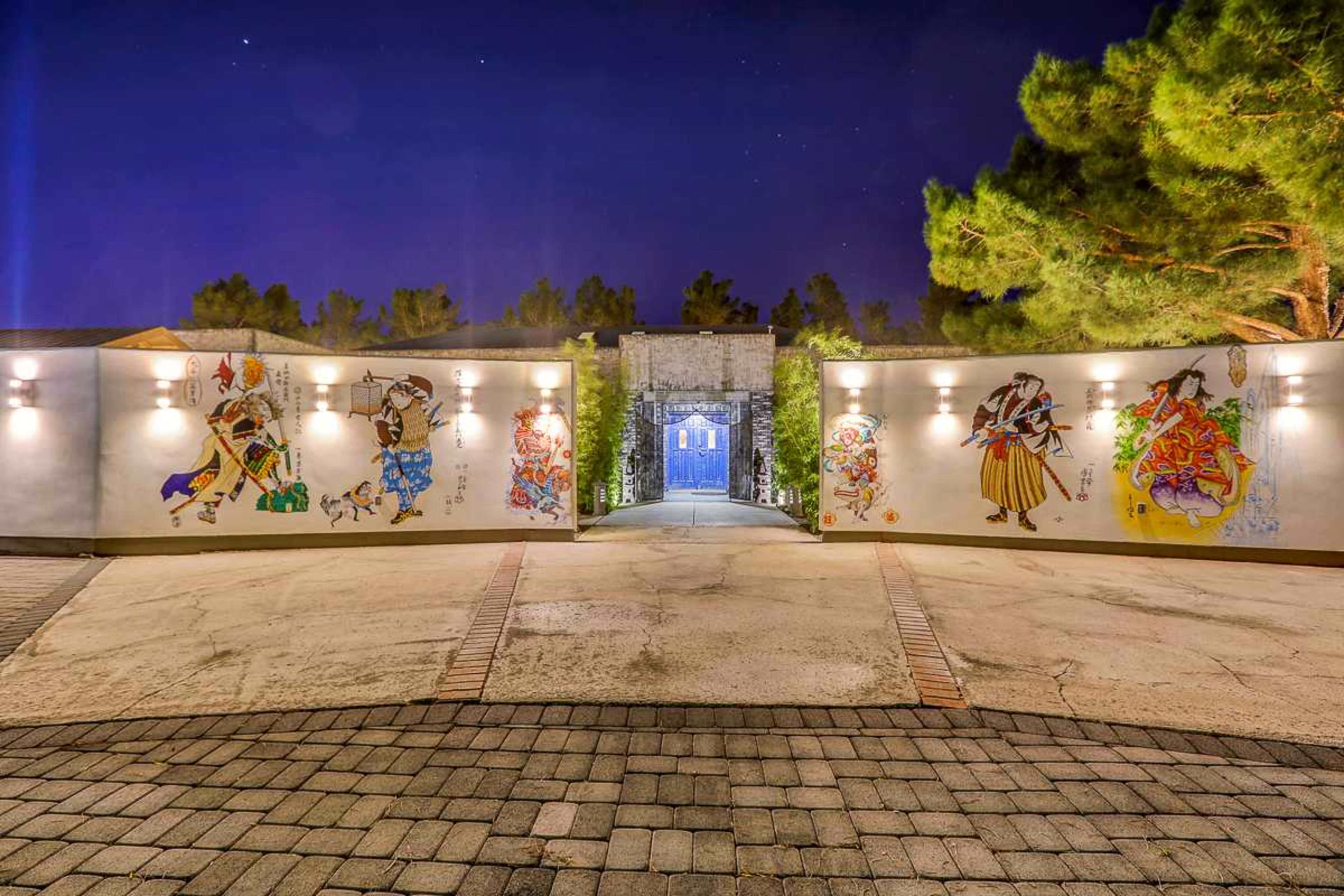 A well-lit entryway with colorful murals on the walls, leading to a blue door surrounded by trees at night.