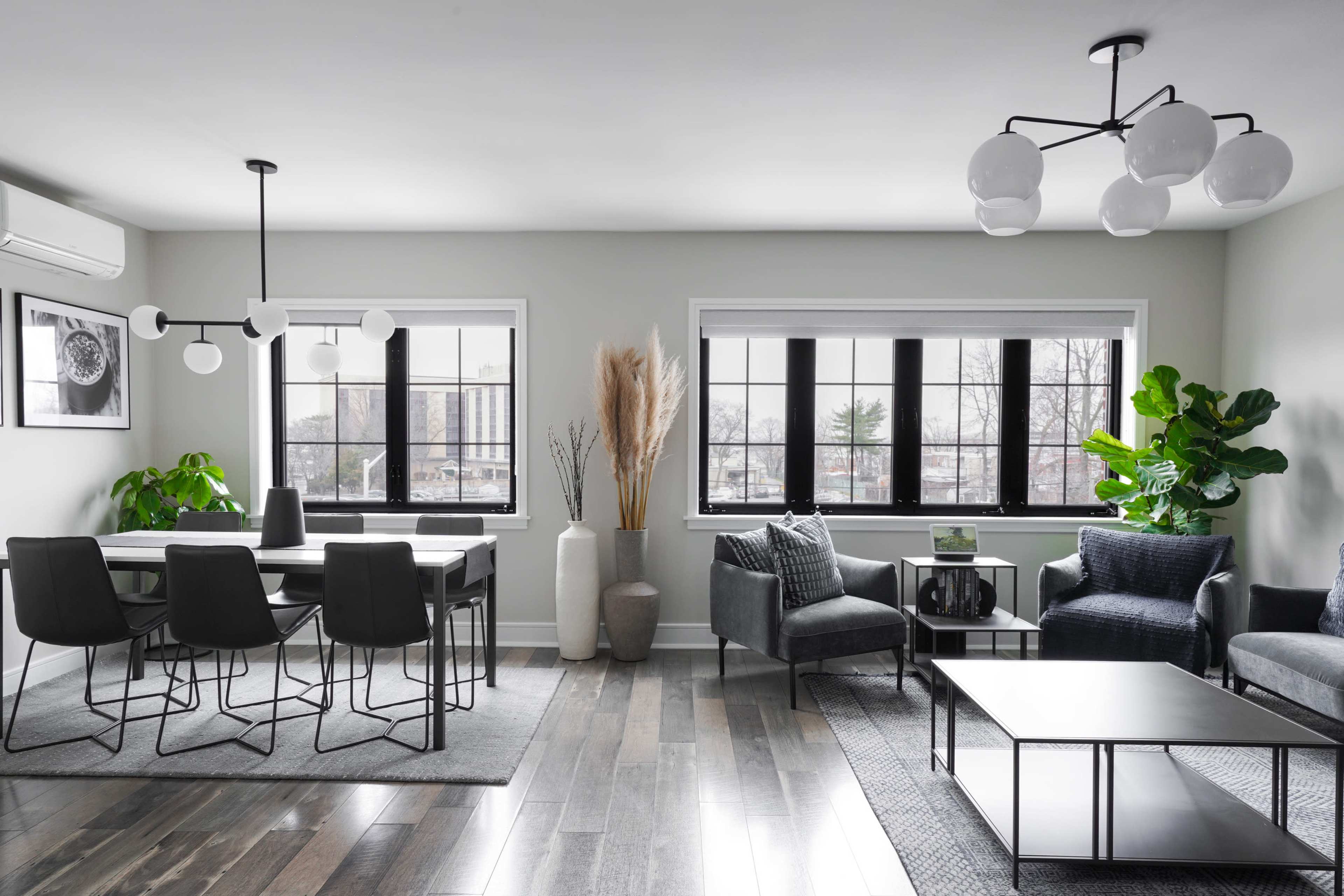 The image shows a modern living room and dining area with large windows, a dining table, and various plants.