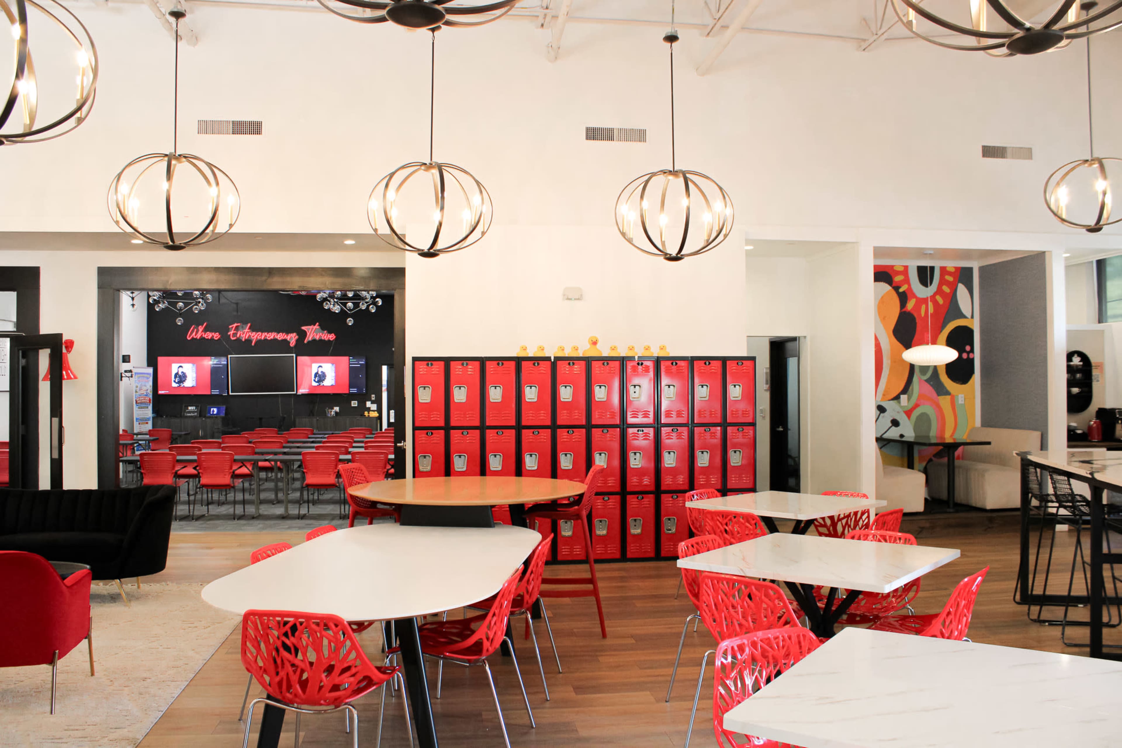 The image shows a modern café or collaborative space featuring red and white furniture, a seating area with circular tables, and a brightly colored wall with lockers in the background.
