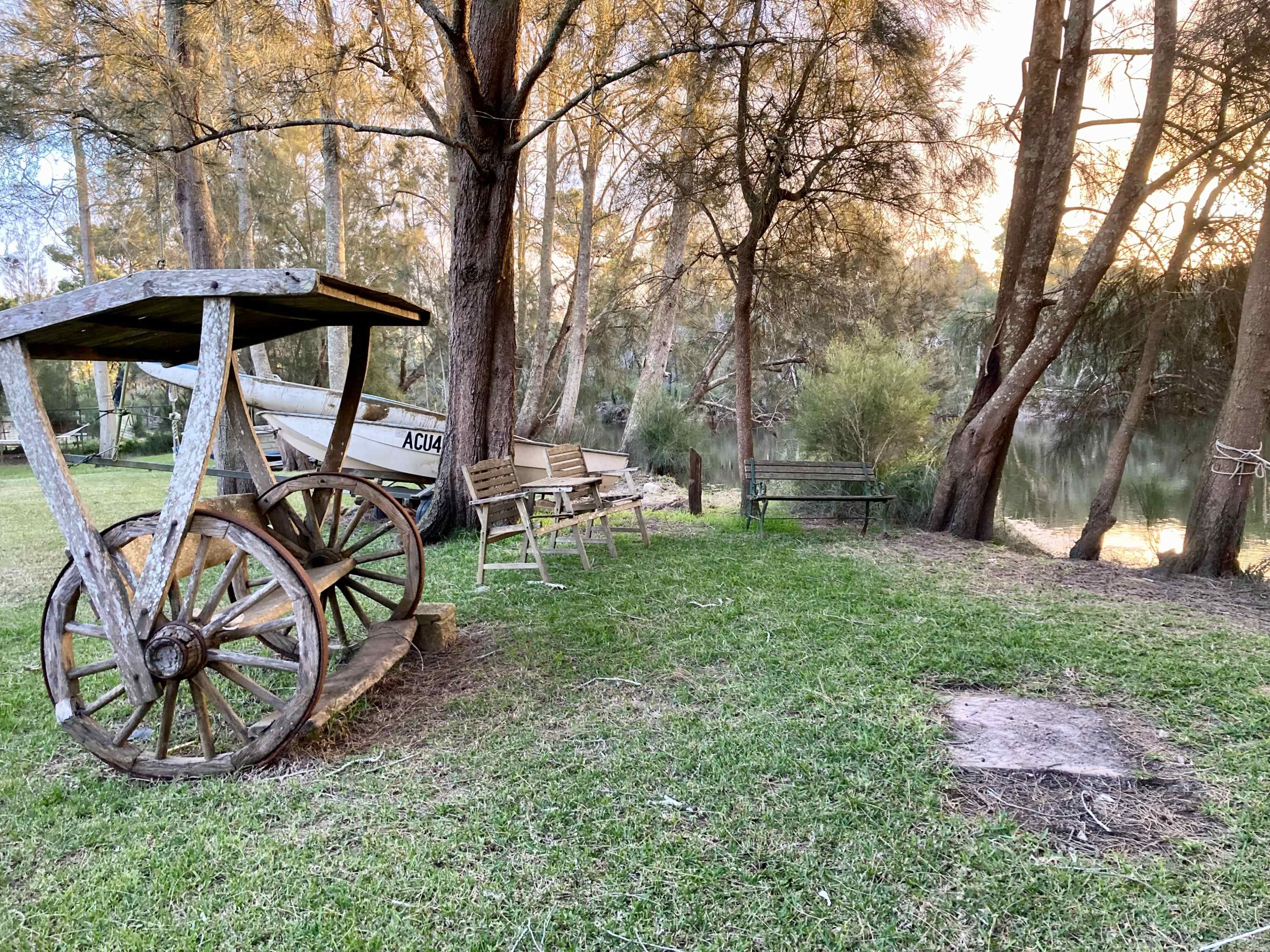 A wooden wagon sits on grass next to several benches beside a river, surrounded by trees.