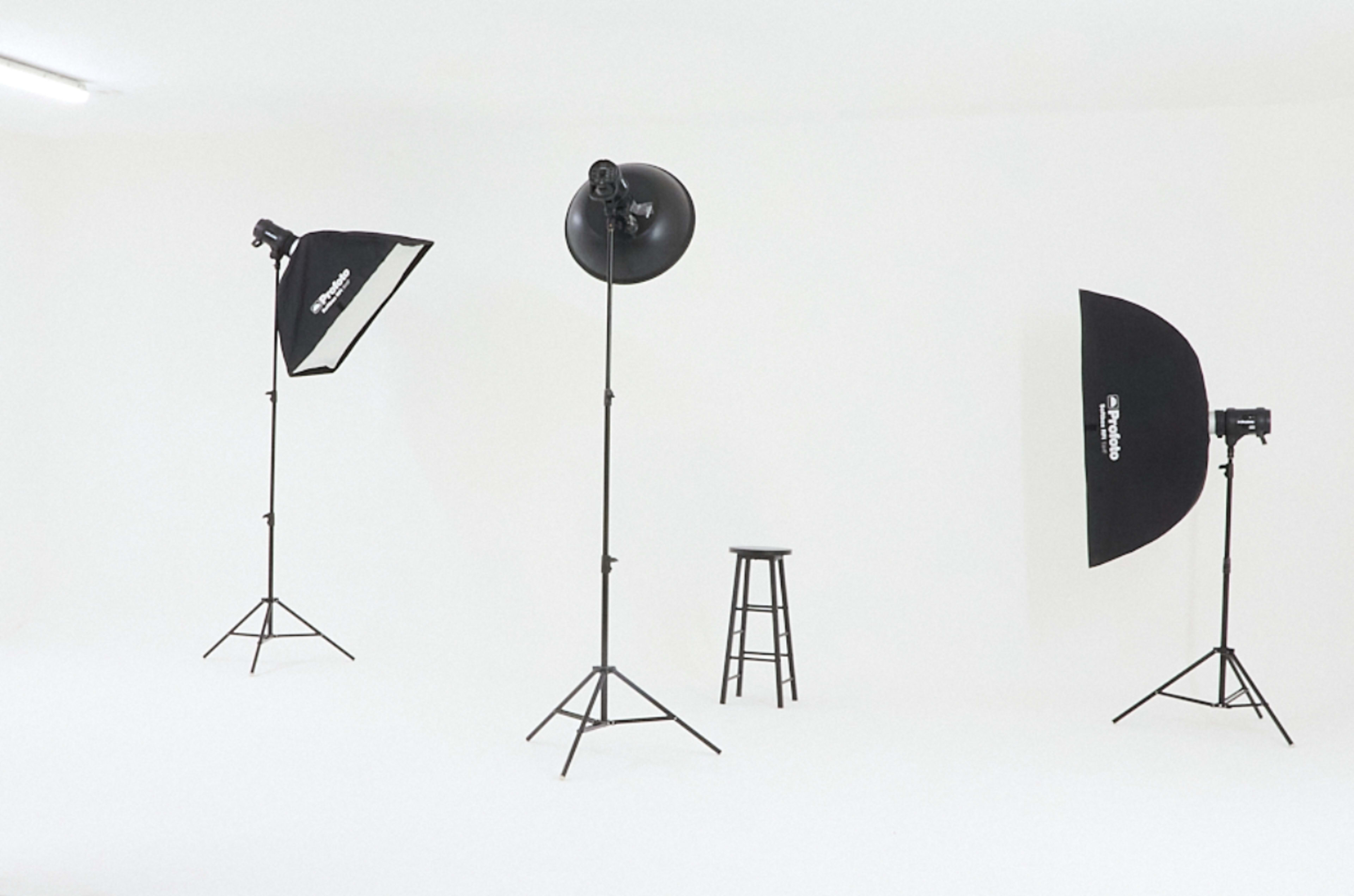 The image shows a minimalist photography studio with two softbox lights, a spotlight, and a black stool on a white background.