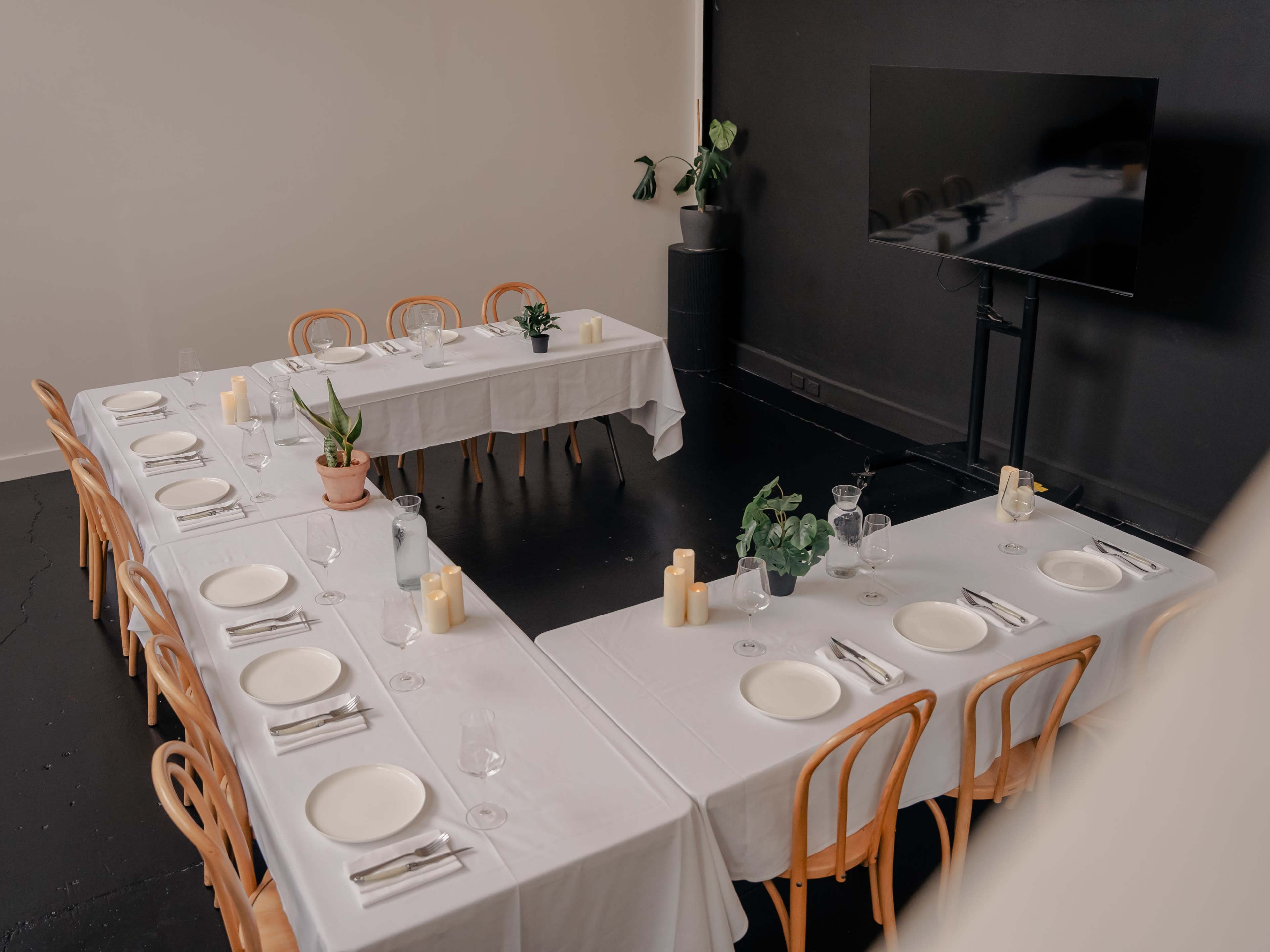 A rectangular dining setup features white tablecloths, plates, glassware, and candles, arranged in a modern space with a large television and potted plants nearby.