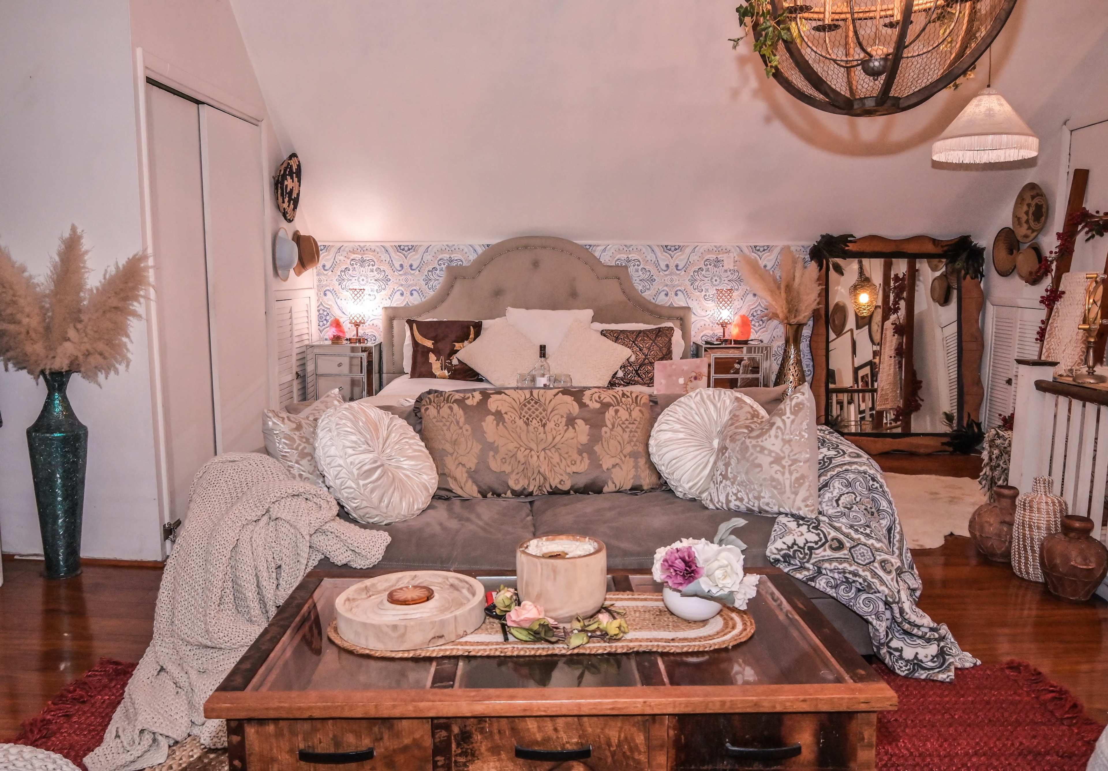 The image shows a cozy bedroom with a large bed adorned with decorative pillows, a wooden coffee table in front, and light-colored walls featuring intricate patterns.