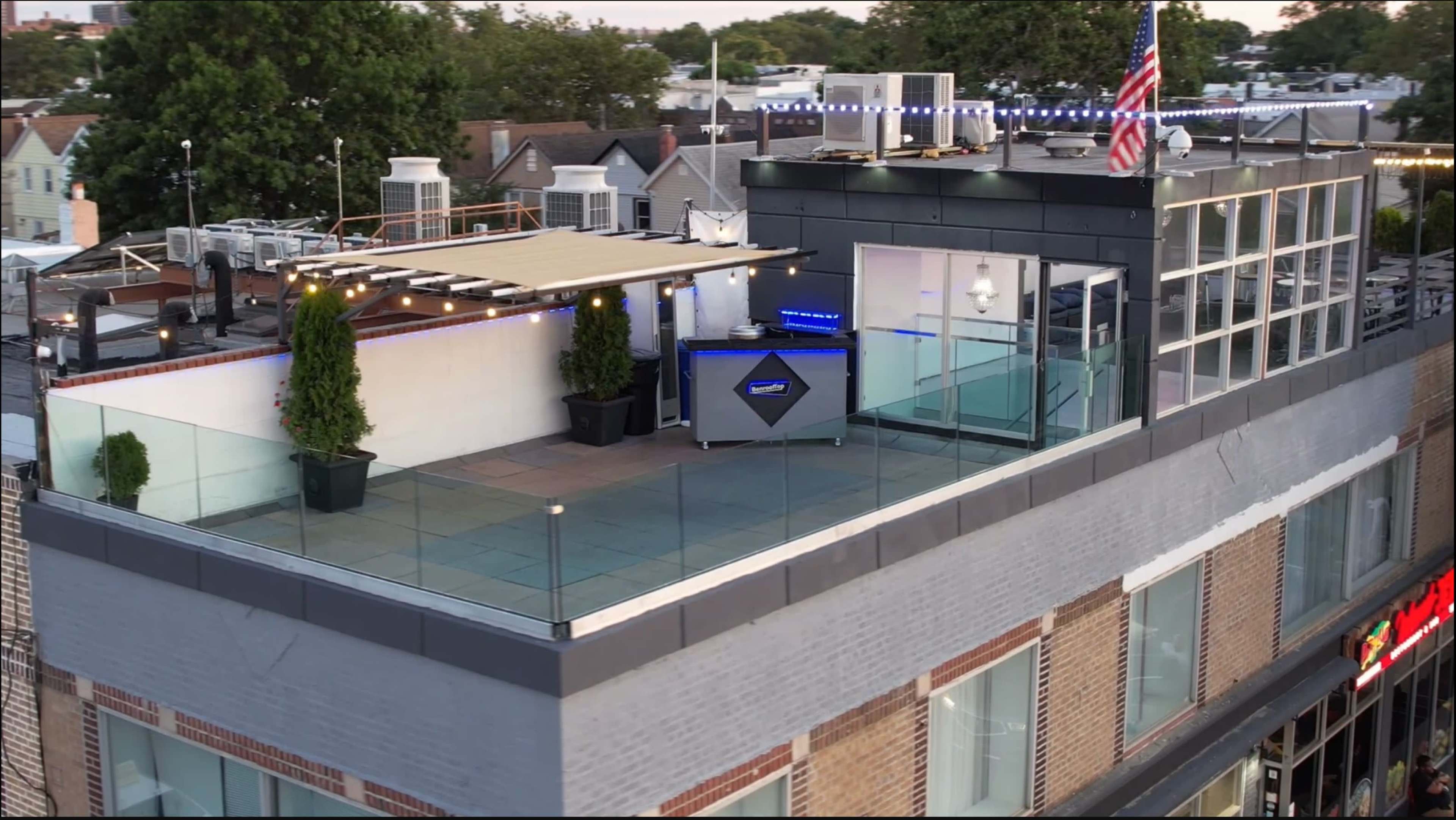 The image shows a rooftop terrace with glass railings, two potted plants, and a bar area, alongside an American flag and various structures in the background.