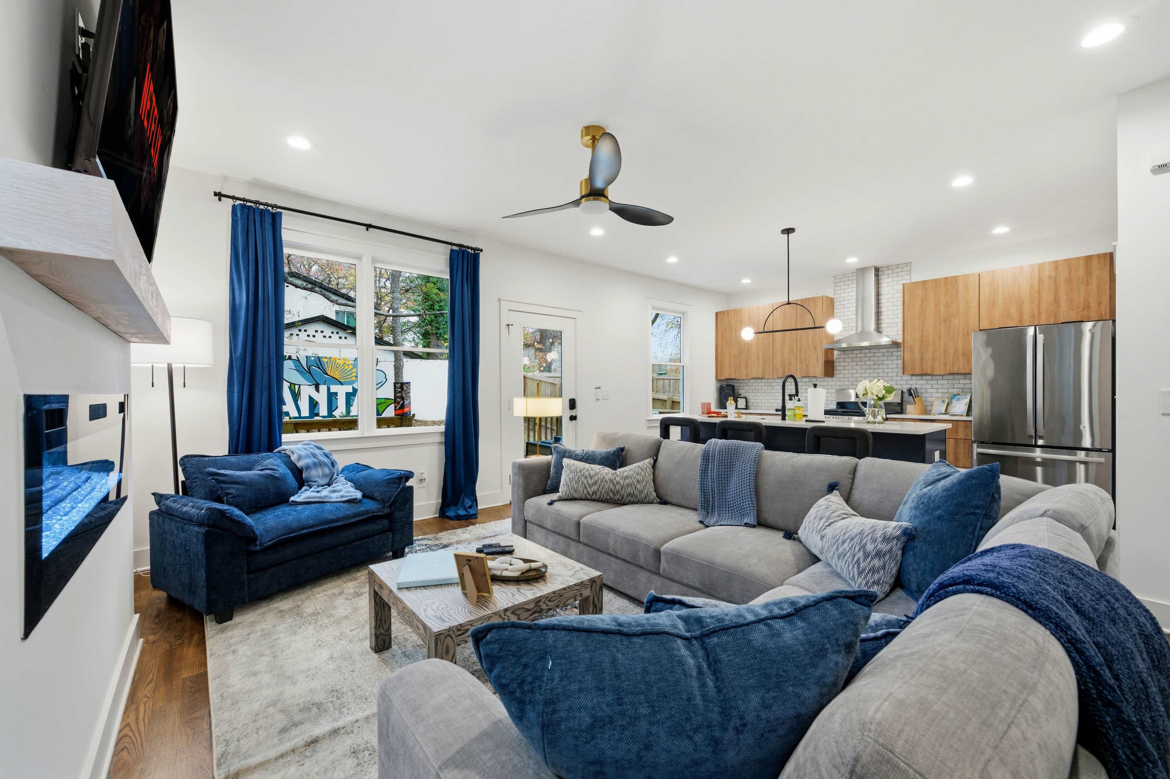 The image shows a modern living room with a large gray sectional sofa, a glass coffee table, a wall-mounted television, and a kitchen area in the background featuring wooden cabinets and a large window.