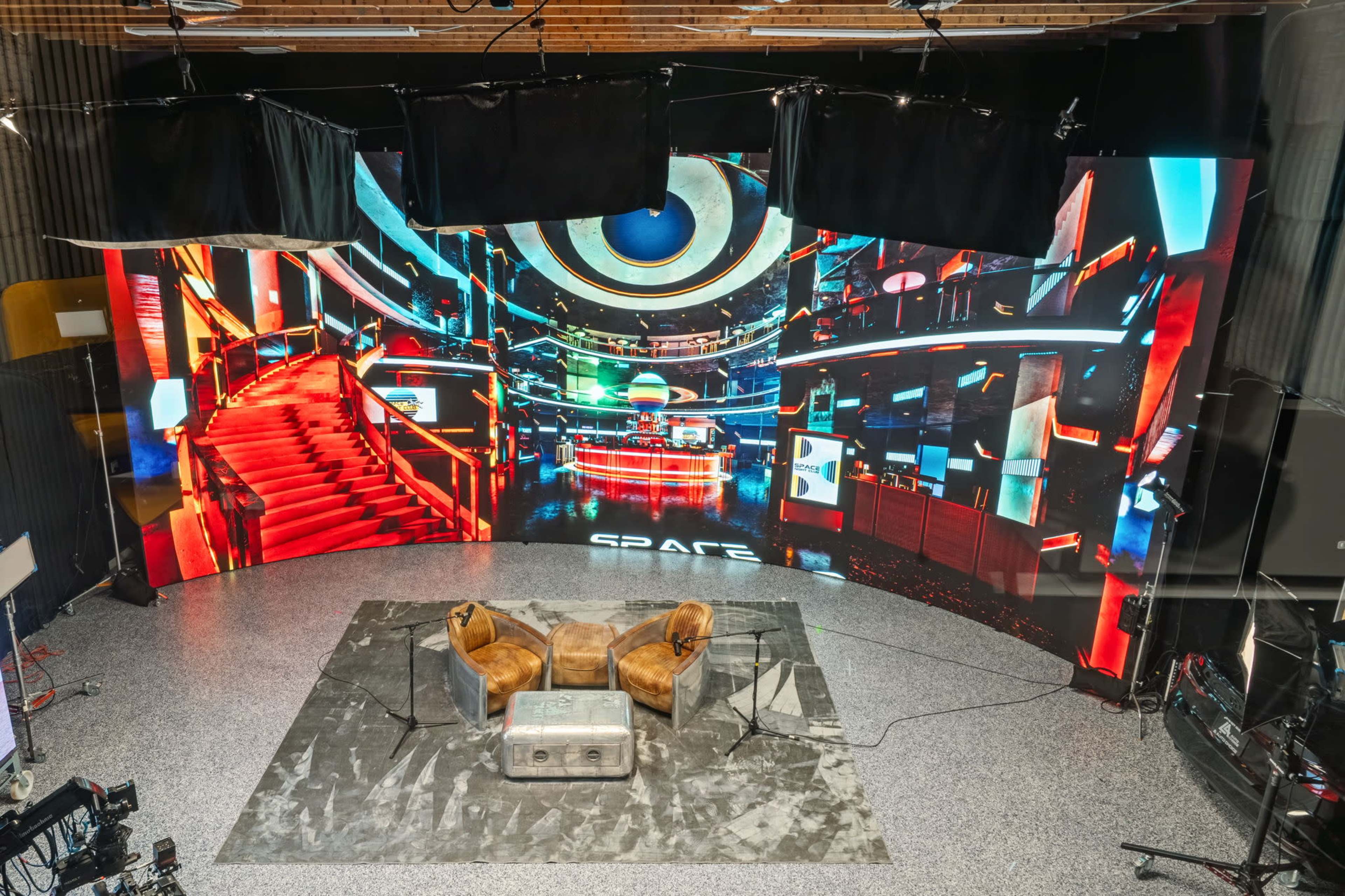 The image shows a modern studio setup with a large digital backdrop featuring vibrant visuals, two brown chairs, and a metallic table on a patterned floor.