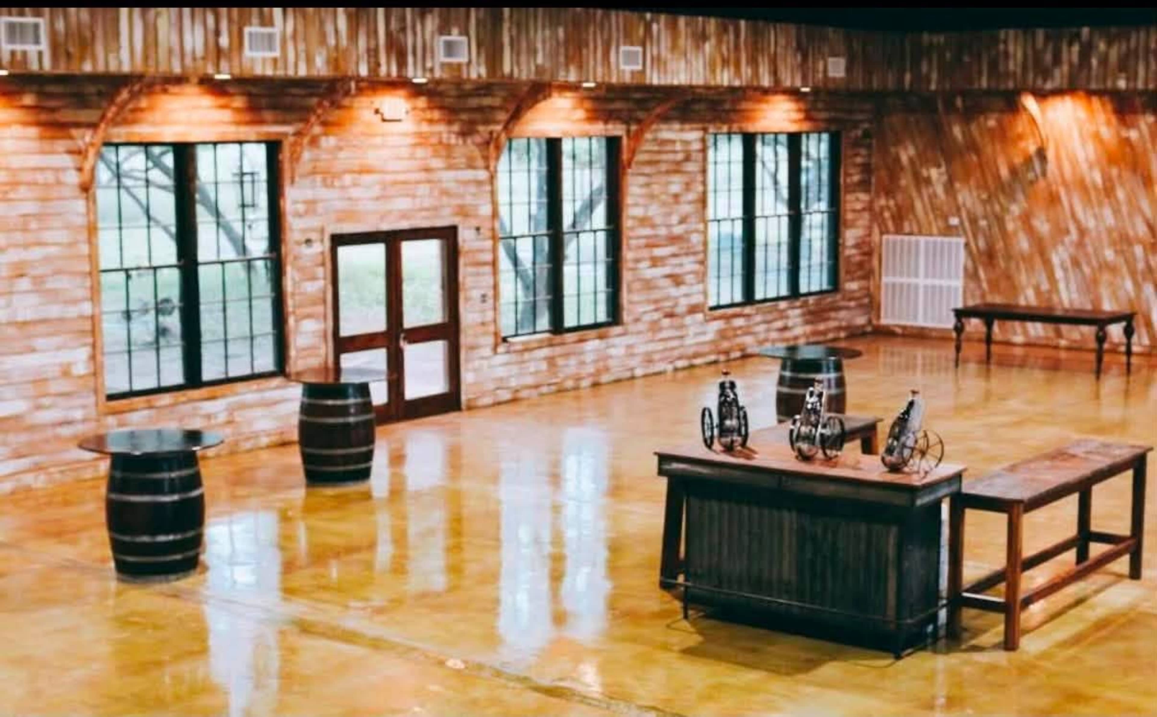 The interior of a rustic venue features wooden walls, large windows, and polished concrete flooring, with a central table and decorative barrels.