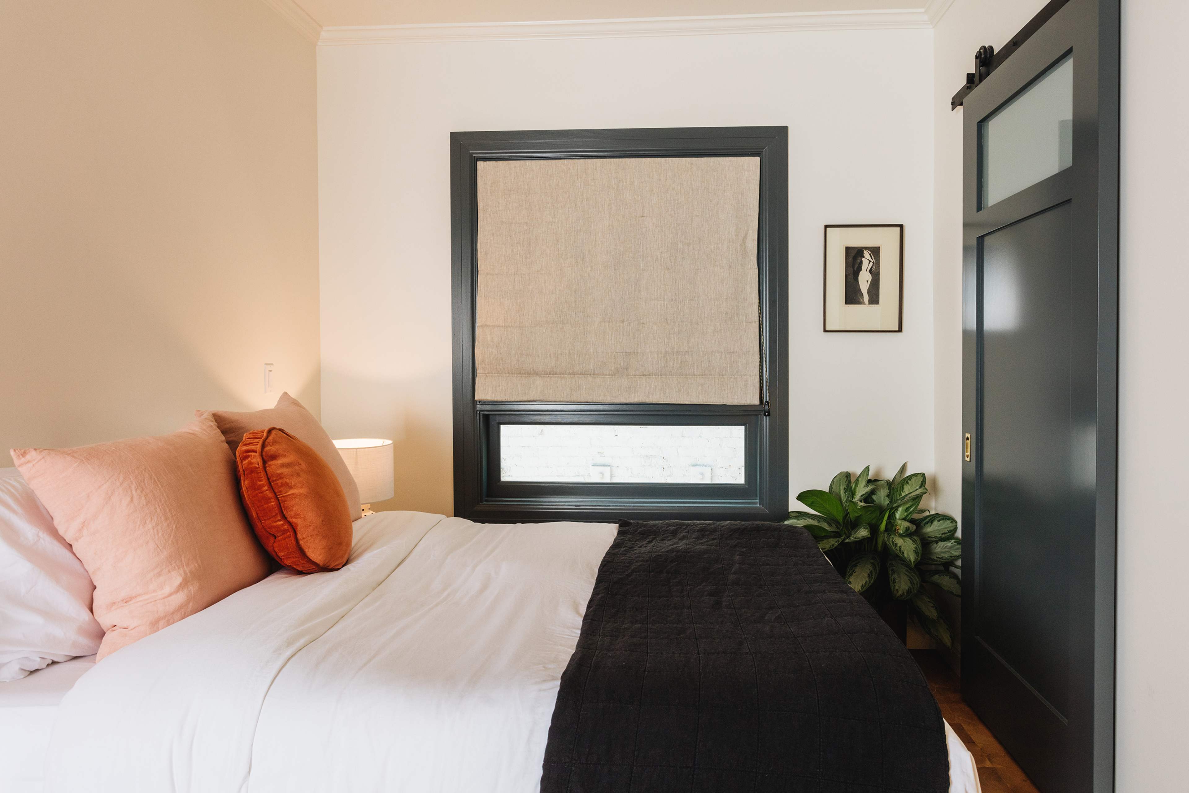 A well-decorated bedroom featuring a bed with white linens and decorative pillows, a bedside lamp, a potted plant, and a window with a shade.