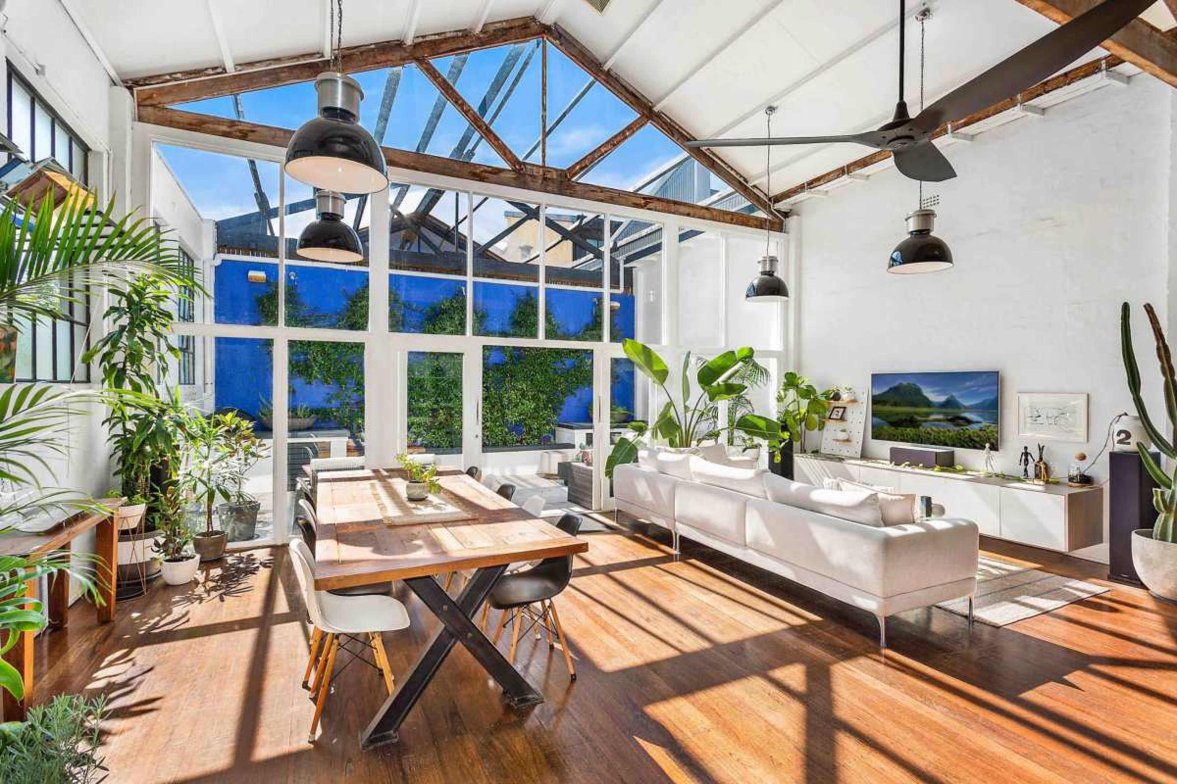 The image shows a bright, airy living space with large windows, wooden beams, and a long dining table surrounded by modern furniture and plants.
