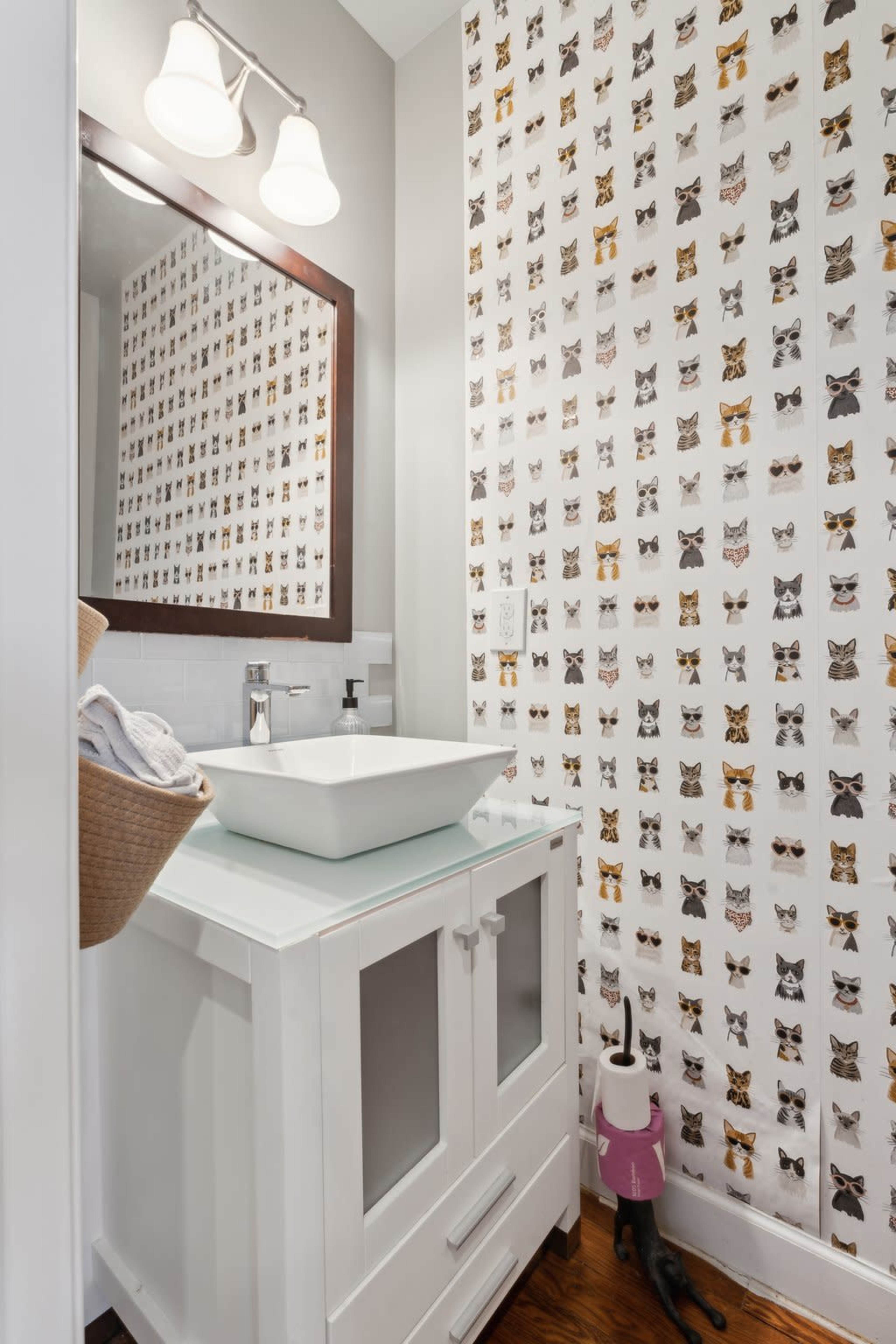The bathroom features a modern white vanity with a rectangular sink, a mirror above it, and a wall decorated with a patterned wallpaper of various cats.