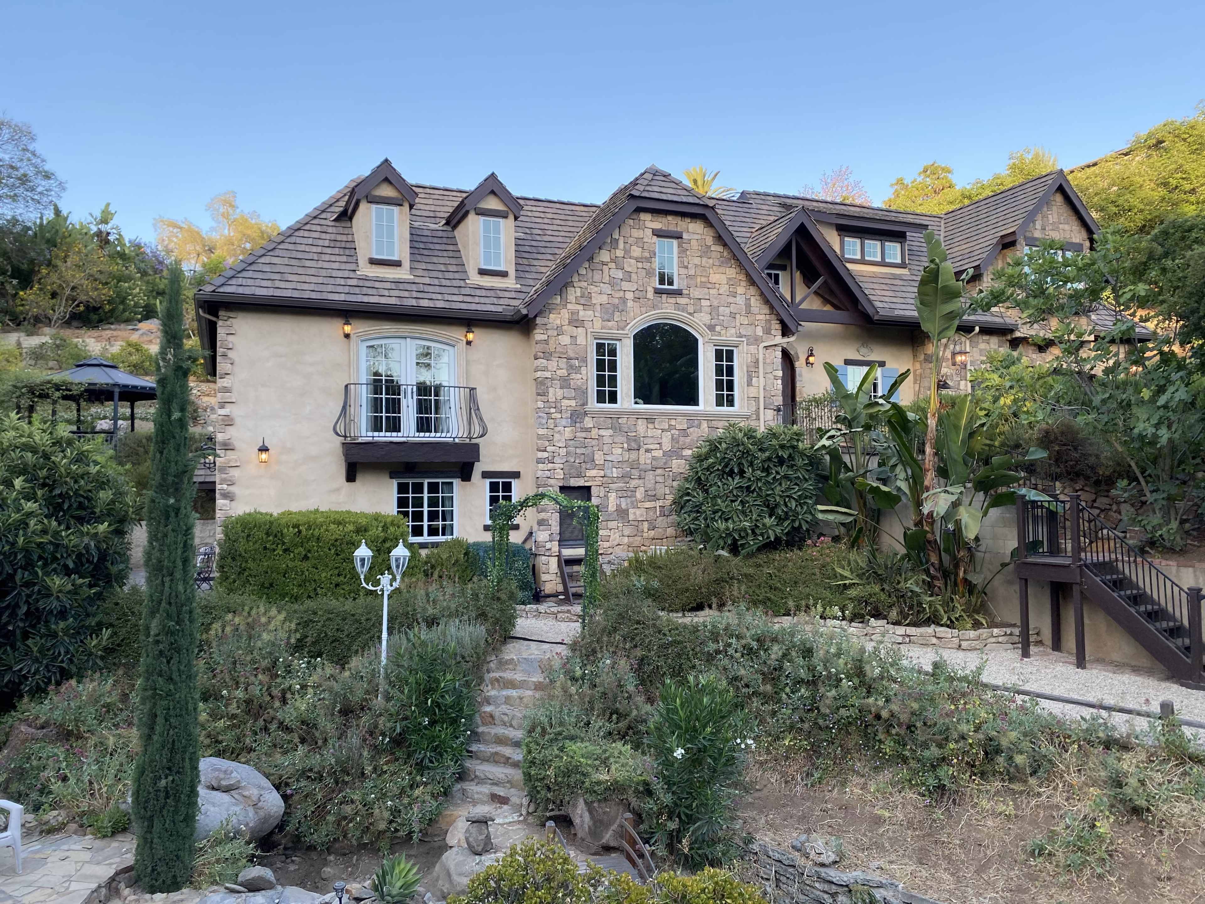 A large, two-story house with a stone facade, surrounded by greenery and featuring a staircase leading to the entrance.